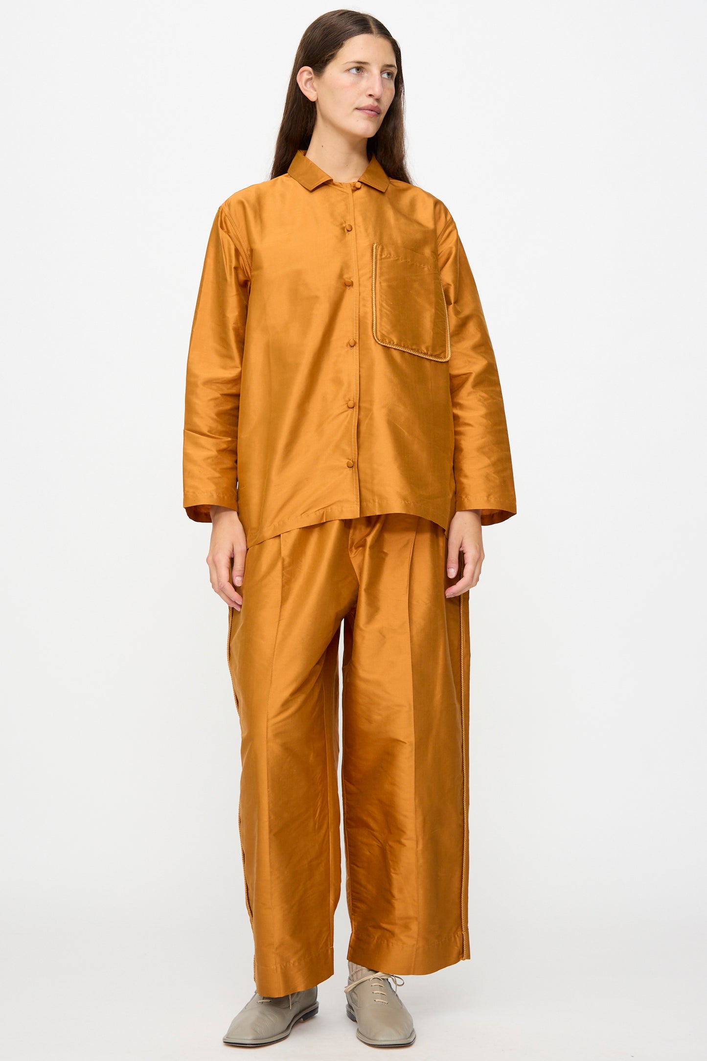 A person stands wearing Cawley’s Silk Dupion Luna Trouser with Trim in Sienna, paired with a loose-fitting, shiny silk dupion copper-colored button-up shirt featuring a chest pocket. They have long hair and are wearing beige shoes.