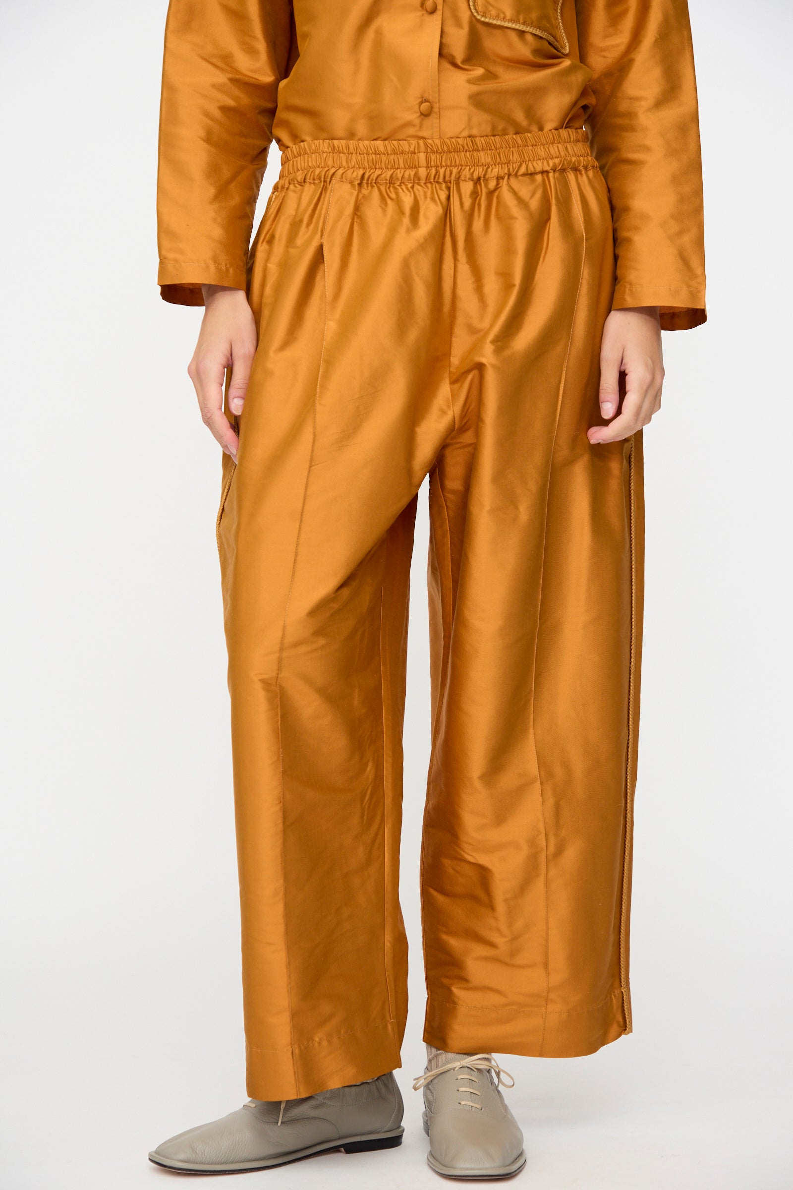 A person wearing the Cawley Silk Dupion Luna Trouser with Trim in Sienna, paired with a matching cropped top and beige lace-up shoes, standing against a plain white background.