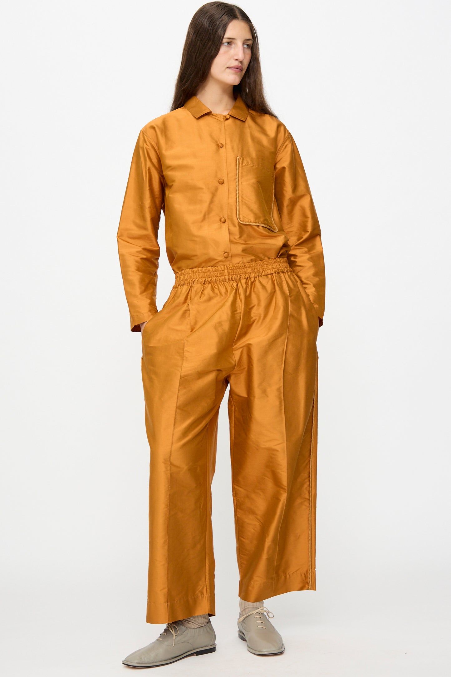 A person stands against a white background wearing Cawley’s Silk Dupion Luna Trouser with Trim in Sienna, styled with a shiny orange silk dupion cropped button-up shirt and light gray shoes.