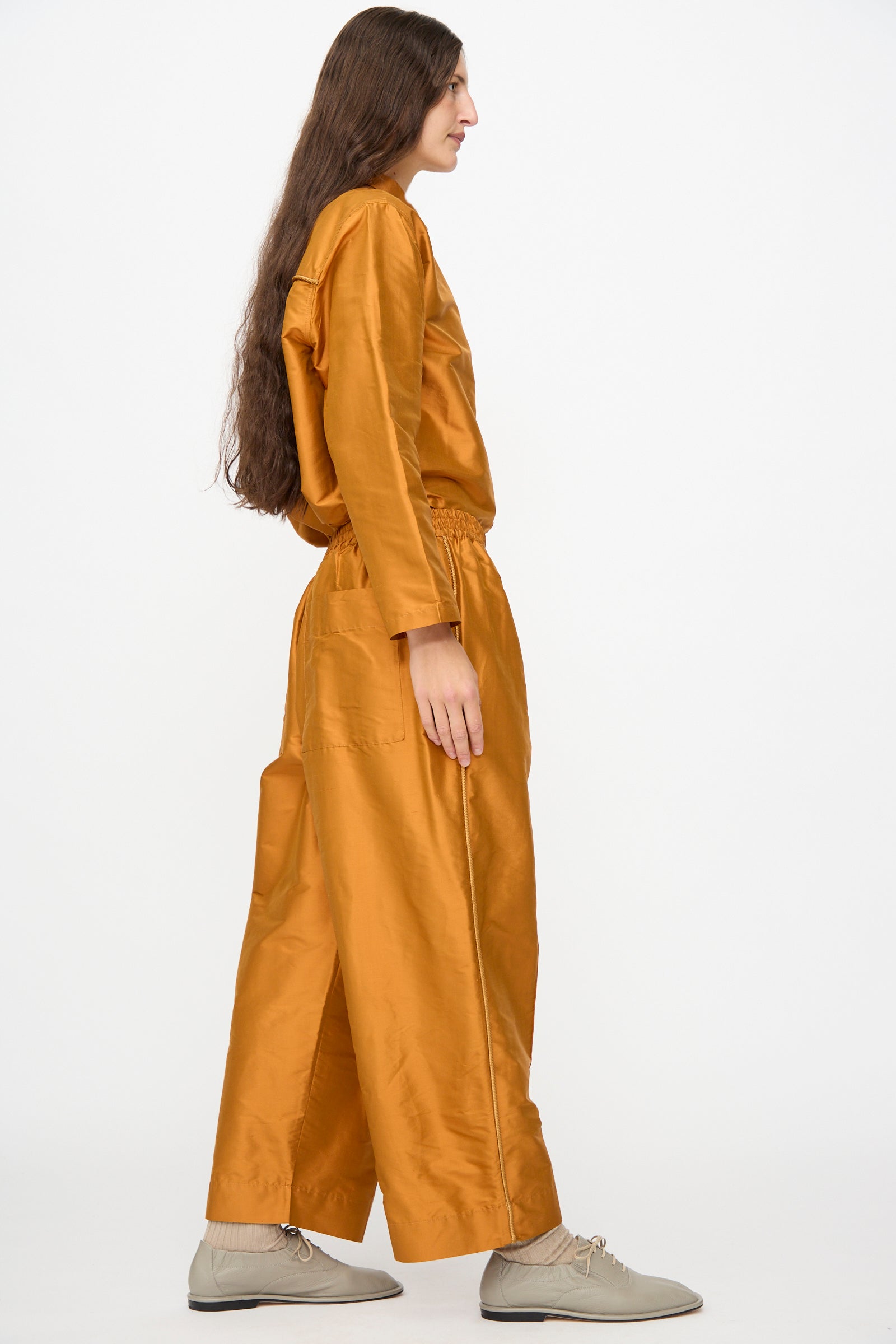 A person with long brown hair, seen in profile, wears the Cawley Silk Dupion Luna Trouser with Trim in Sienna, paired with a loose-fitting, shiny silk orange top and beige lace-up shoes, standing against a plain white background.