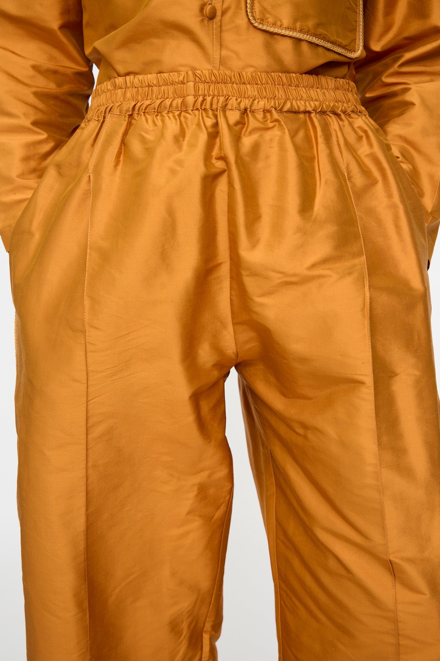 A person wearing the Cawley Silk Dupion Luna Trouser with Trim in Sienna—a pair of shiny orange-brown silk dupion pants featuring an elastic waistband—paired with a matching top, stands with hands in pockets, shown from waist to mid-thigh.
