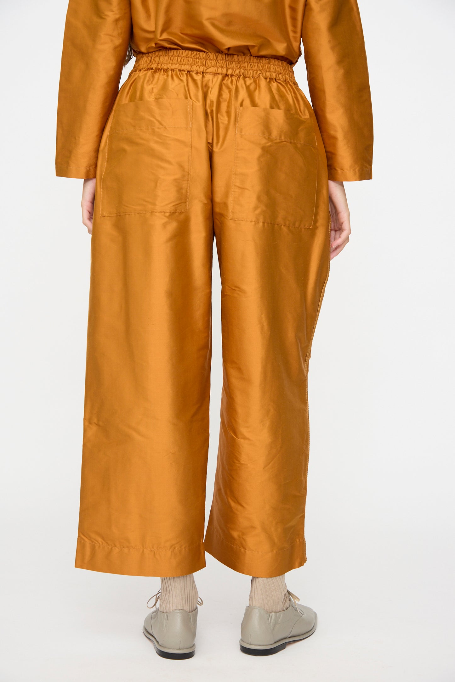 A person is photographed from the back against a plain white background wearing the Cawley Silk Dupion Luna Trouser with Trim in Sienna, featuring a loose fit, cropped length, and an elastic waistband. The outfit is styled with beige socks and light gray shoes.