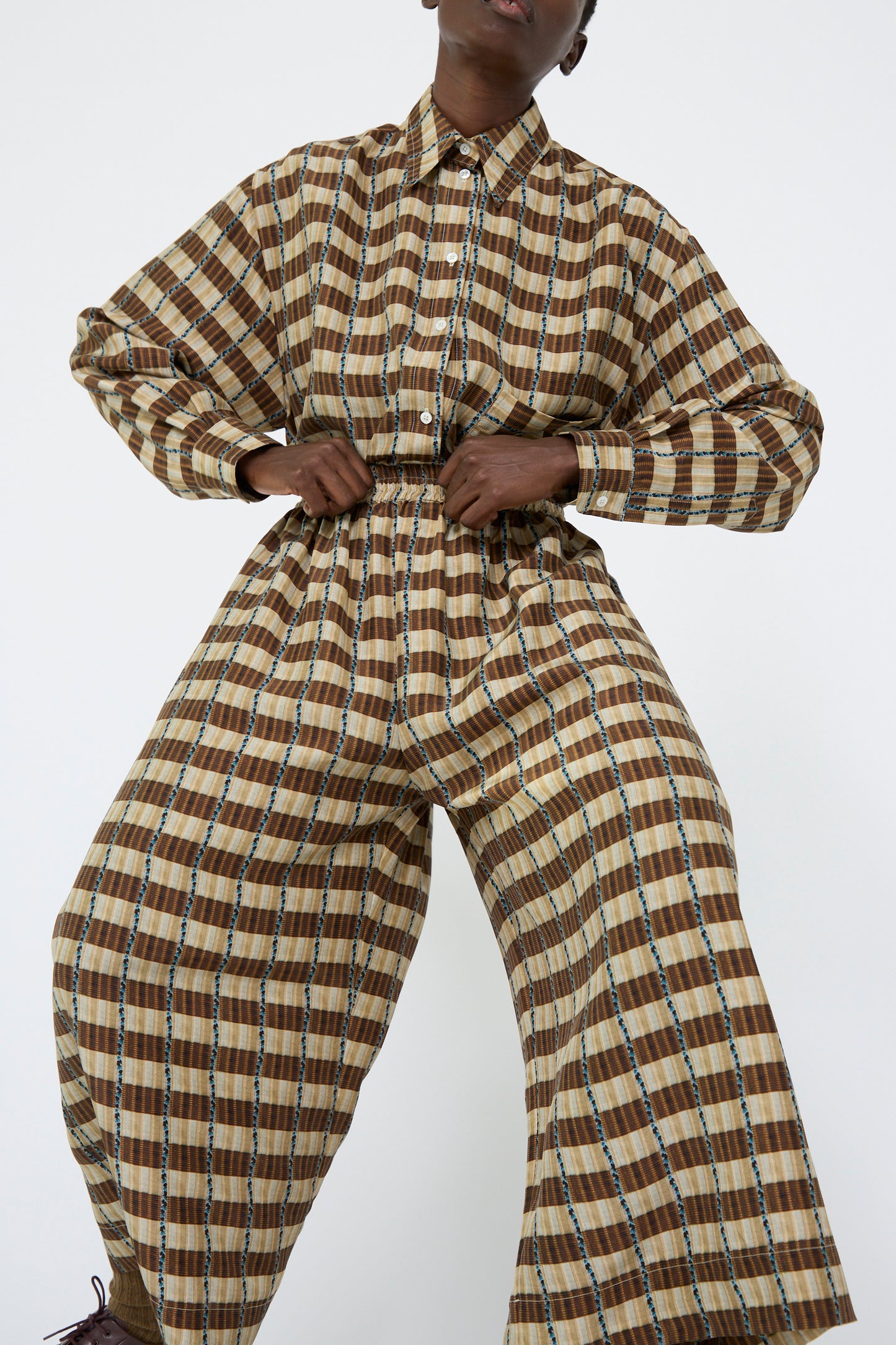 A person strikes a dynamic pose against a plain white background, showcasing Cawley's Silk Printed May Trouser in Brown Plaid. The ensemble's wide-leg pants feature an elasticated waist for comfort and versatility, and relaxed fit.