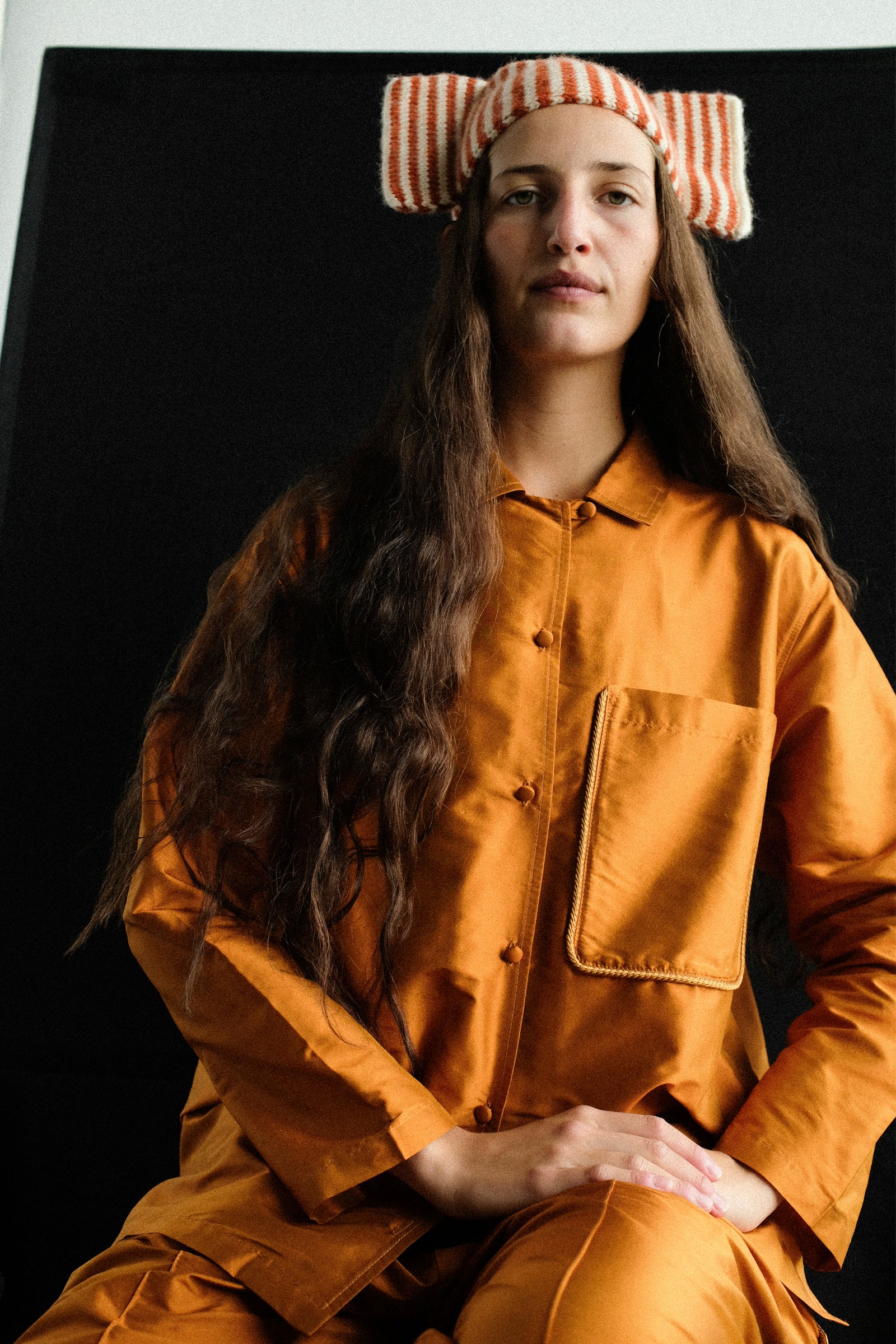 A model wearing Cawley's Brushed British Wool Stripe Bow Hat in Natural and Autumn, and Silk Dupion August Shirt in Sienna, sitting on a stool against a black background.