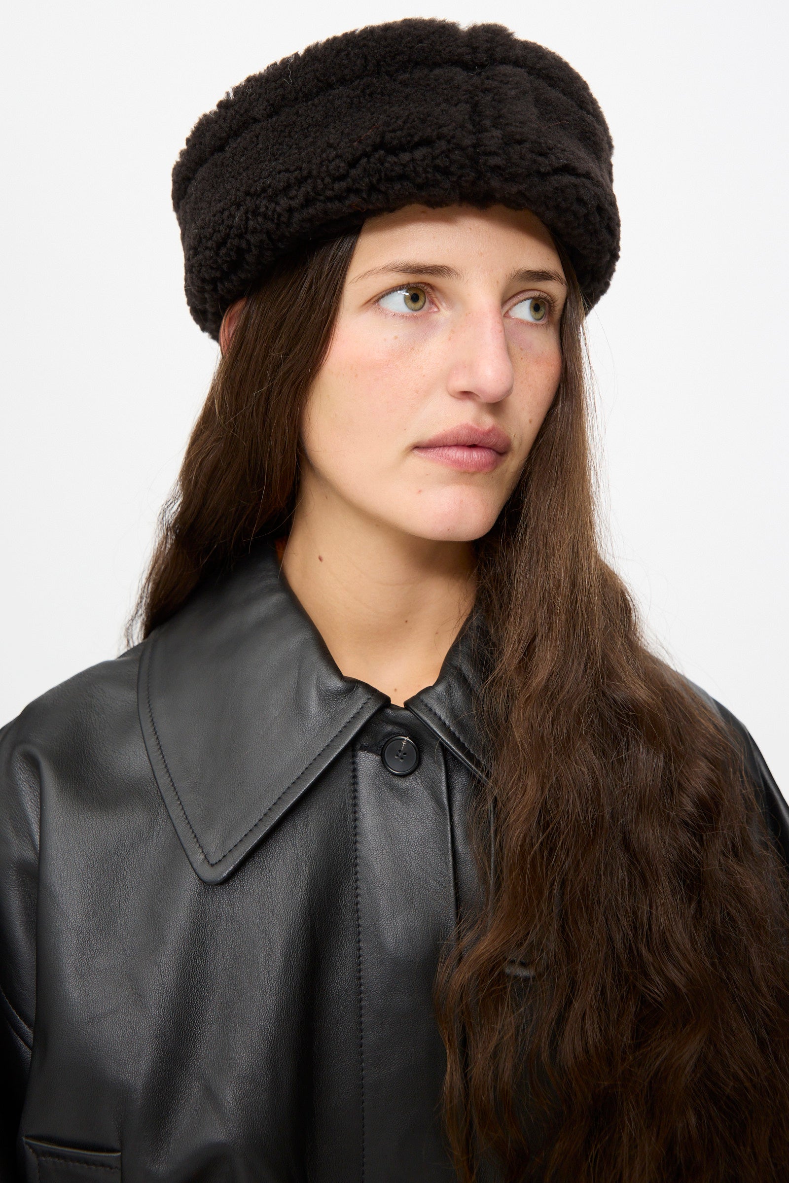 A woman with long brown hair wears the Suede Back Curly Hair Cap in Chocolate by Cawley and a black faux leather coat, looking slightly to her left against a plain white background. 