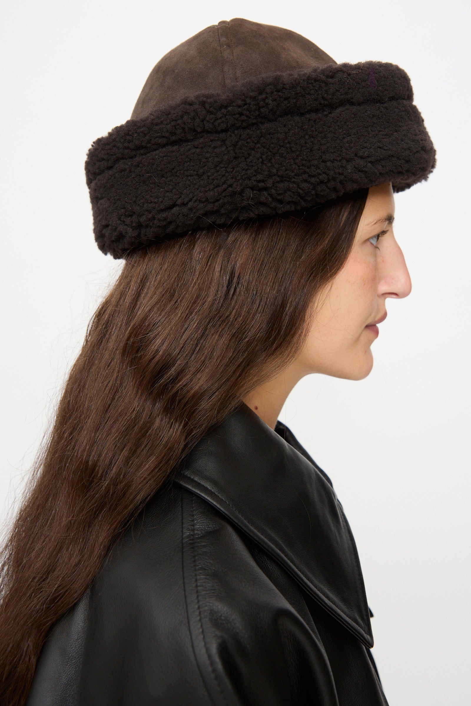 A person in profile wears the Cawley Suede Back Curly Hair Cap in Chocolate and a black leather jacket, with long brown hair visible against a plain white background.