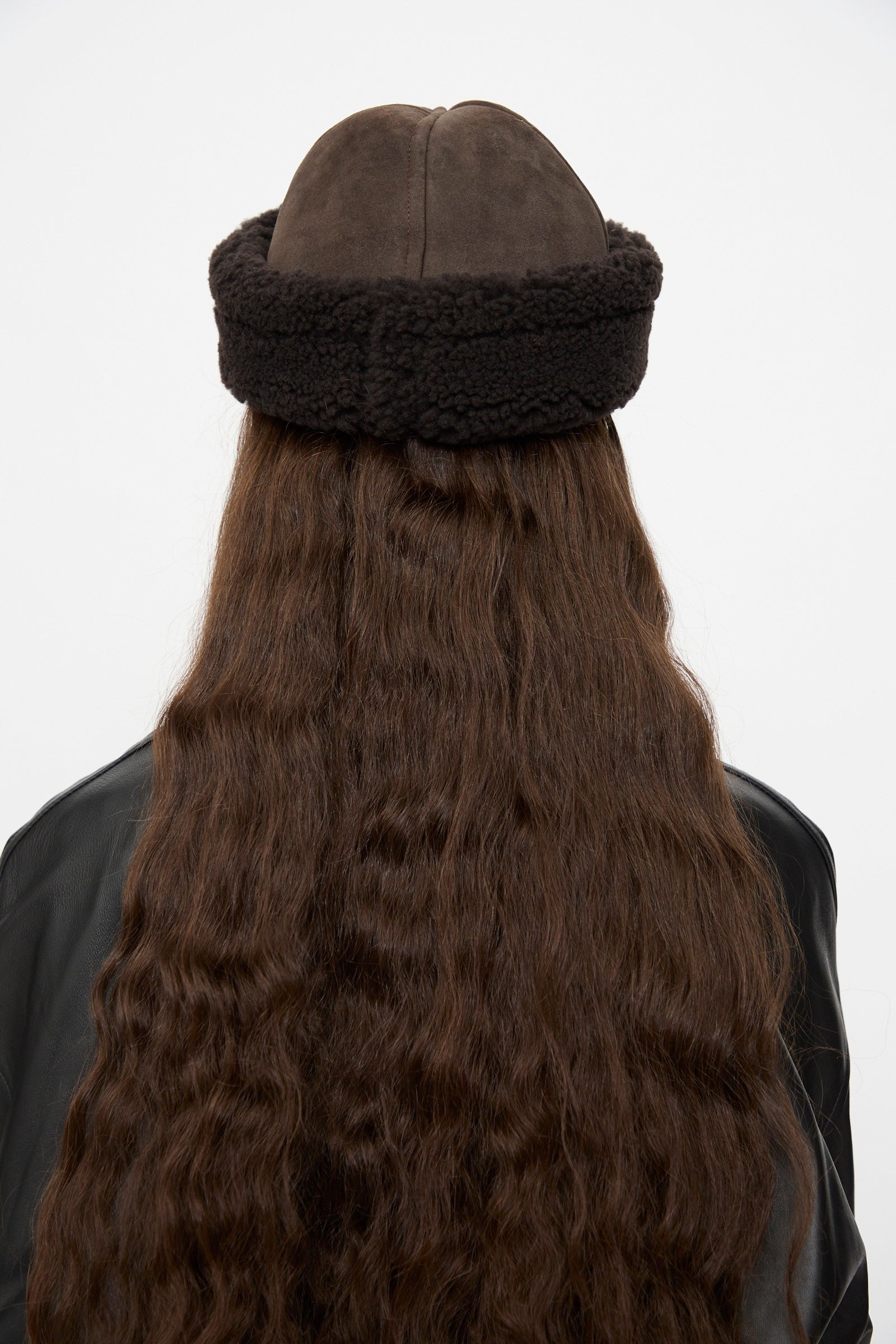 A person with long wavy brown hair, seen from behind against a plain light background, is wearing the Cawley Suede Back Curly Hair Cap in Chocolate and a black leather jacket.