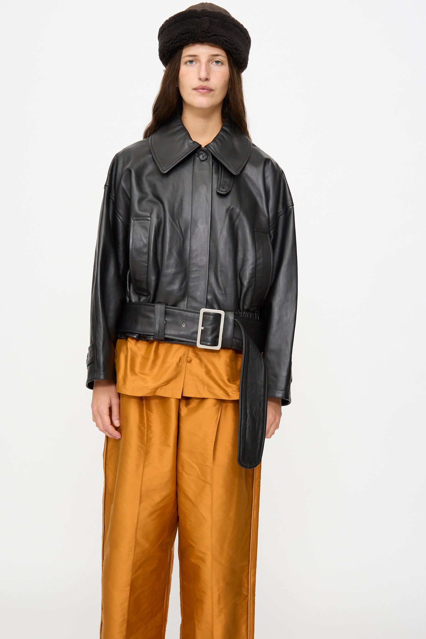 A person wearing a black leather jacket with a belt, gold satin pants, and the Suede Back Curly Hair Cap in Chocolate by Cawley, standing against a plain white background.
