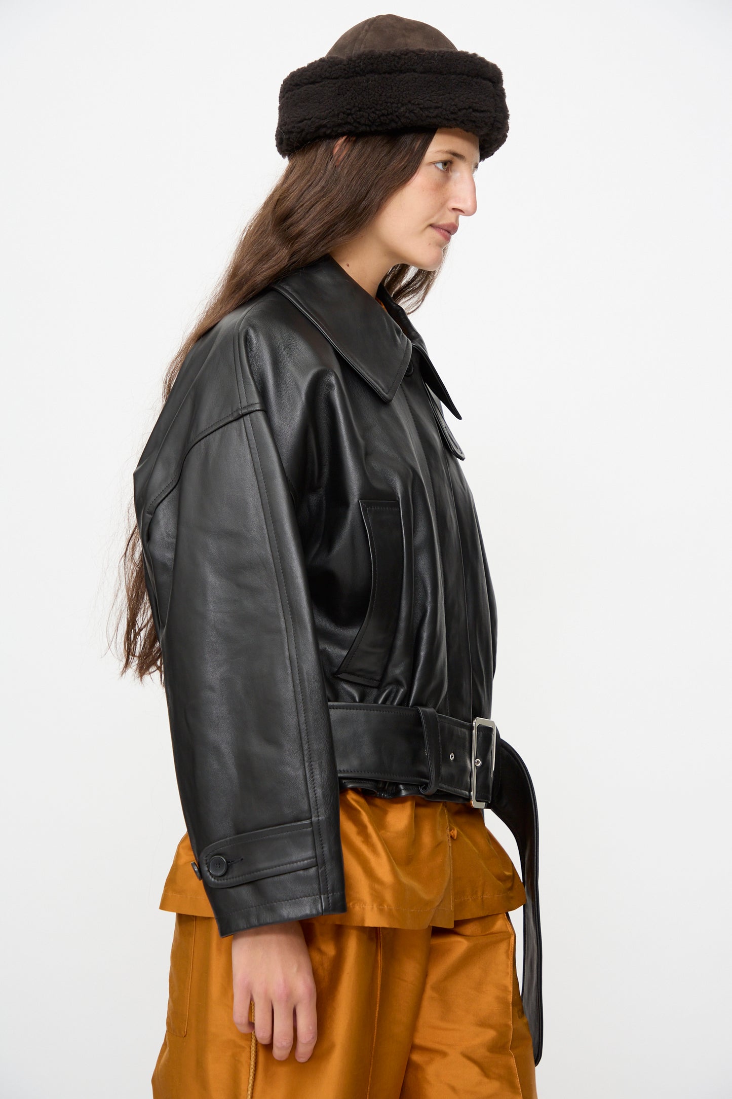 A person in profile view wearing a black leather jacket with belt, brown pants, and the Cawley Suede Back Curly Hair Cap in Chocolate, set against a plain white background.