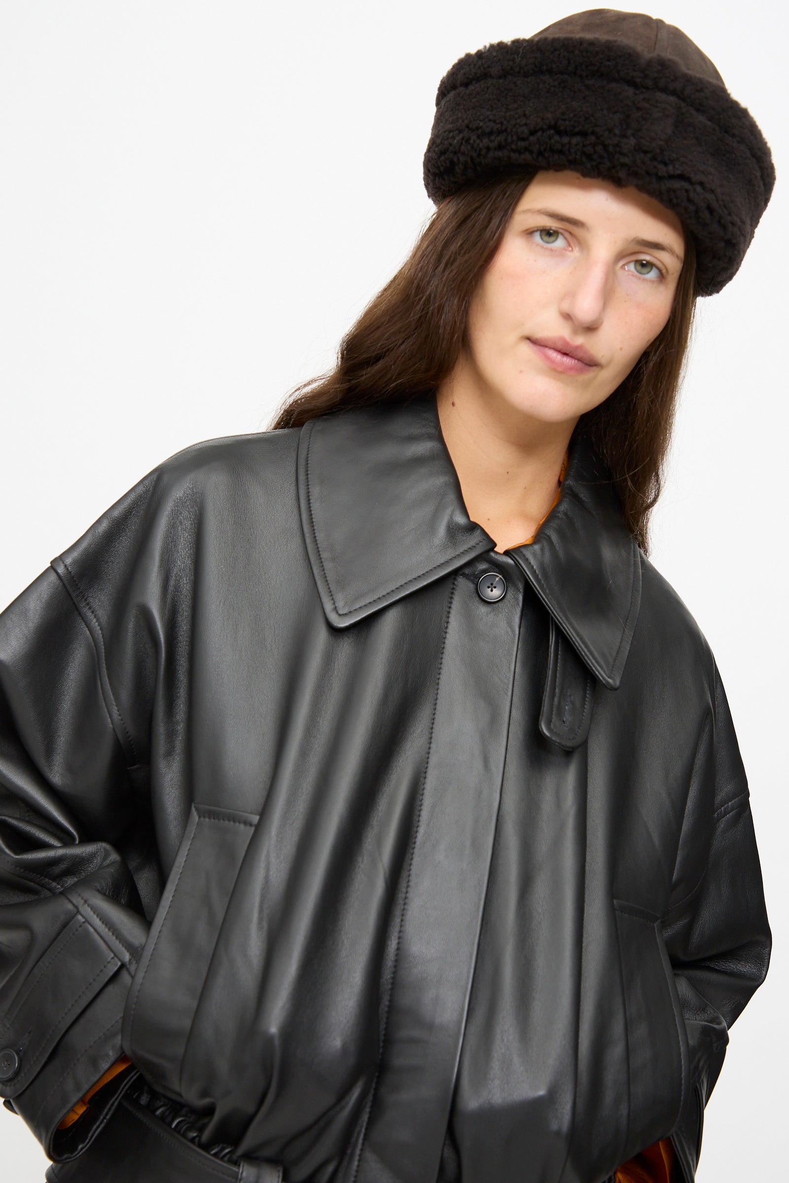 A woman wearing a black leather coat and the Cawley Suede Back Curly Hair Cap in Chocolate stands against a plain white background, looking at the camera.