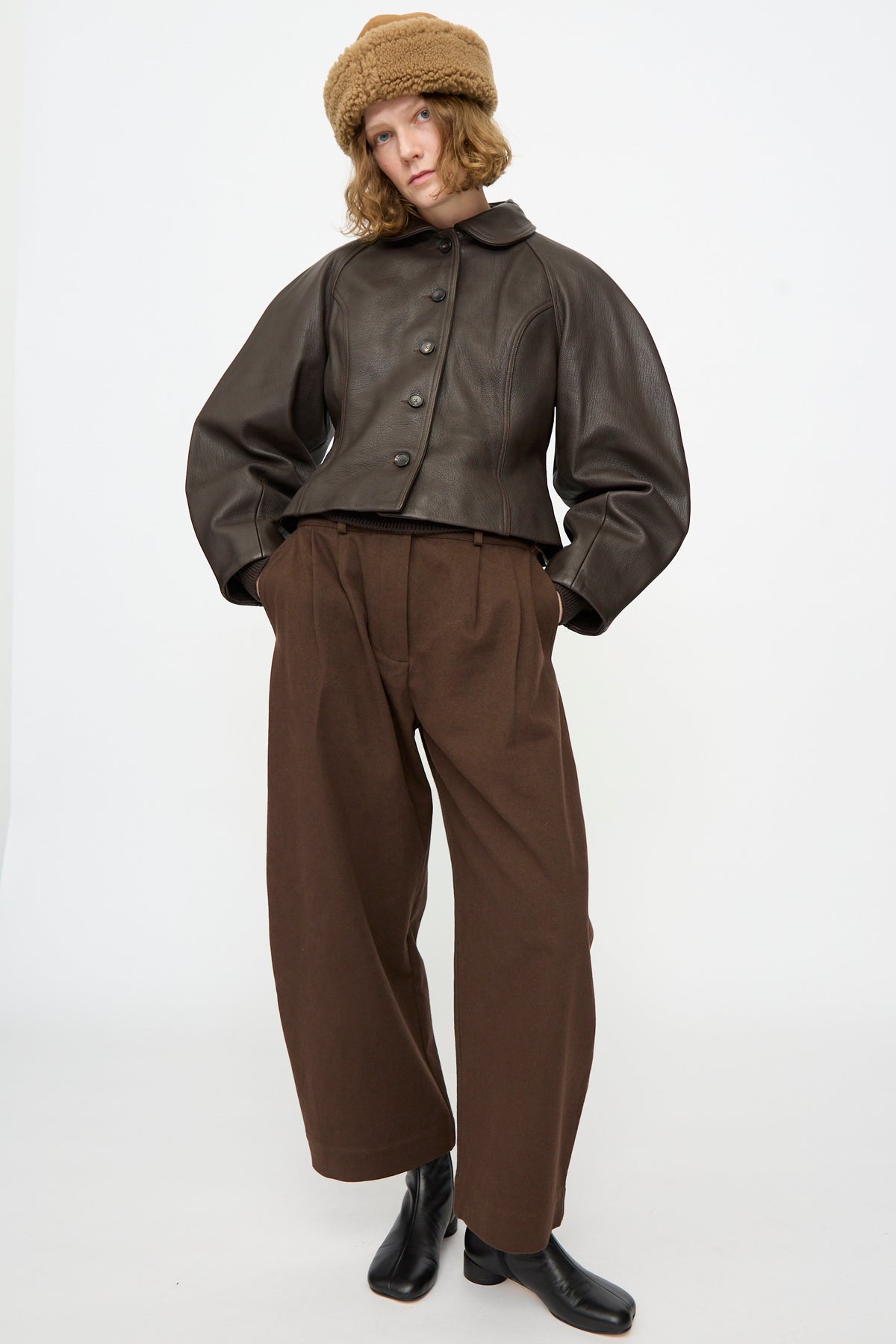 A person wearing a brown leather jacket, wide brown pants, black boots, and the Suede Back Curly Hair Cap in Tan by Cawley stands against a plain white background. This stylish outfit is complemented by the warm cap from Cawley, expertly crafted in London.