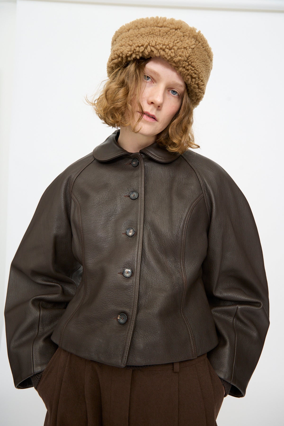 A person wearing a brown leather jacket, brown pants, and the Suede Back Curly Hair Cap in Tan by Cawley stands in front of a plain white background. Crafted in London, this stylish cap showcases classic British craftsmanship and elevates the outfit.