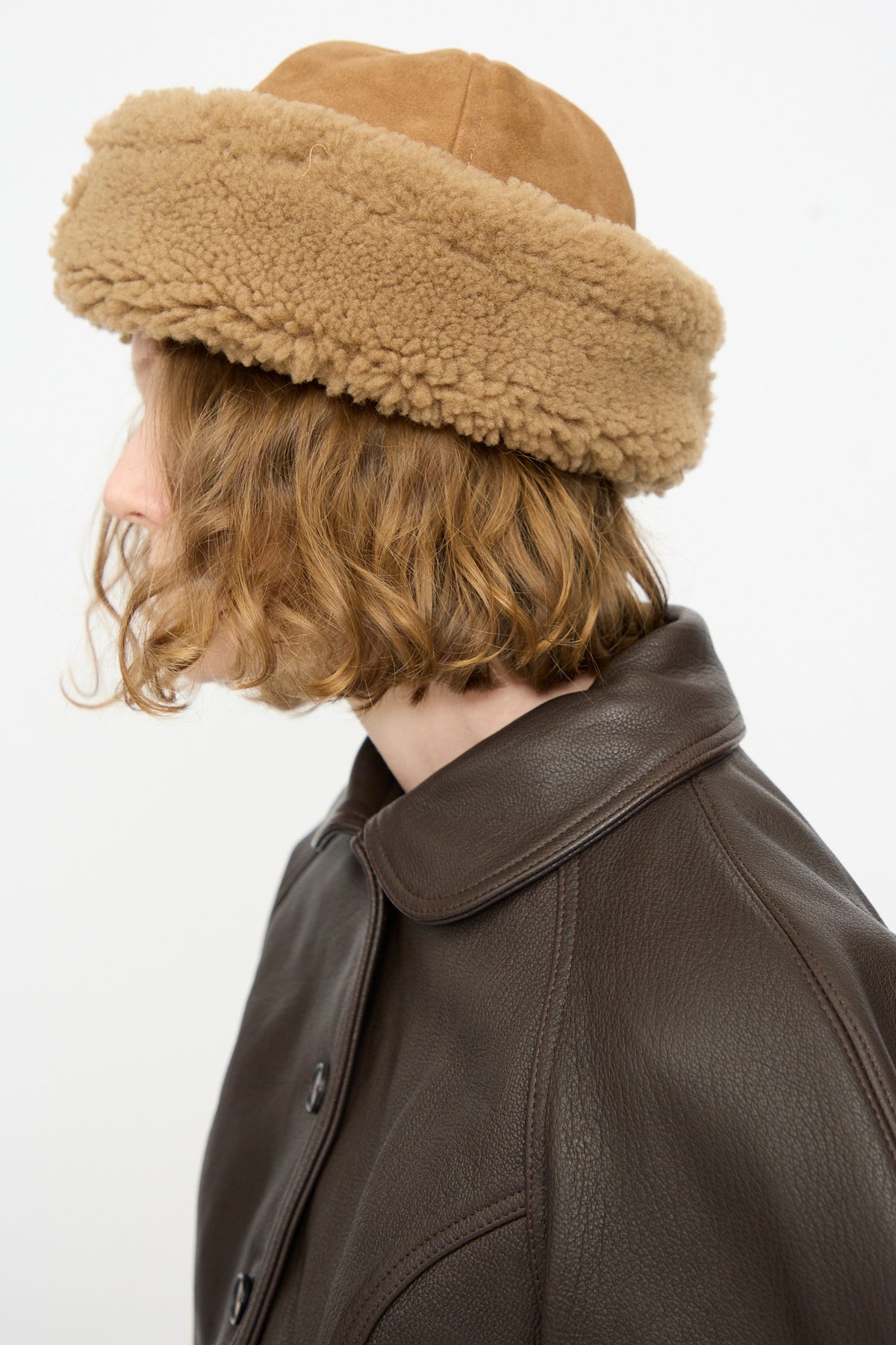 A person with wavy, shoulder-length hair wears the Cawley Suede Back Curly Hair Cap in Tan along with a dark brown leather jacket, facing left against a plain white background.
