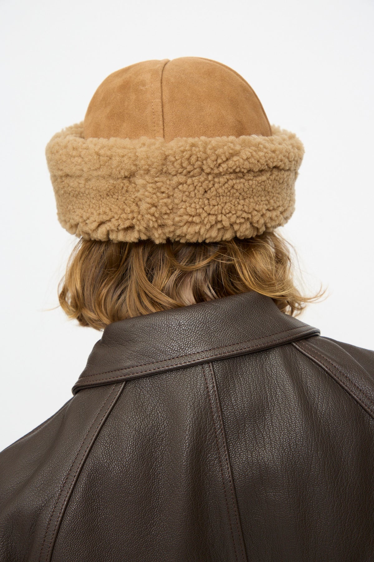 A person with shoulder-length hair, seen from behind against a plain background, is wearing the Cawley Suede Back Curly Hair Cap in Tan and a dark brown leather jacket.
