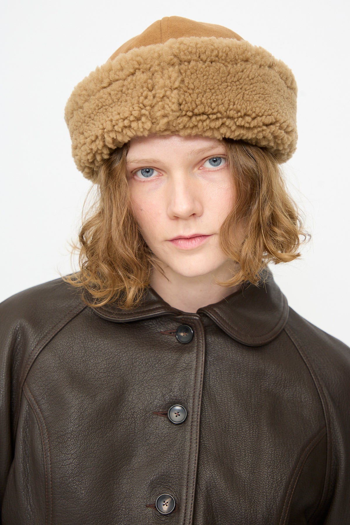 A person with shoulder-length hair wears the Cawley Suede Back Curly Hair Cap in Tan and a dark brown leather coat, facing forward against a plain white background.