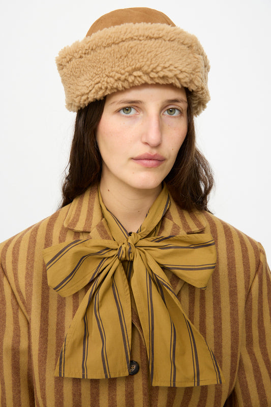 A woman wears the Cawley Suede Back Curly Sheepskin Cap in Tan, paired with a tan and brown striped coat and matching bow, posing against a plain background.