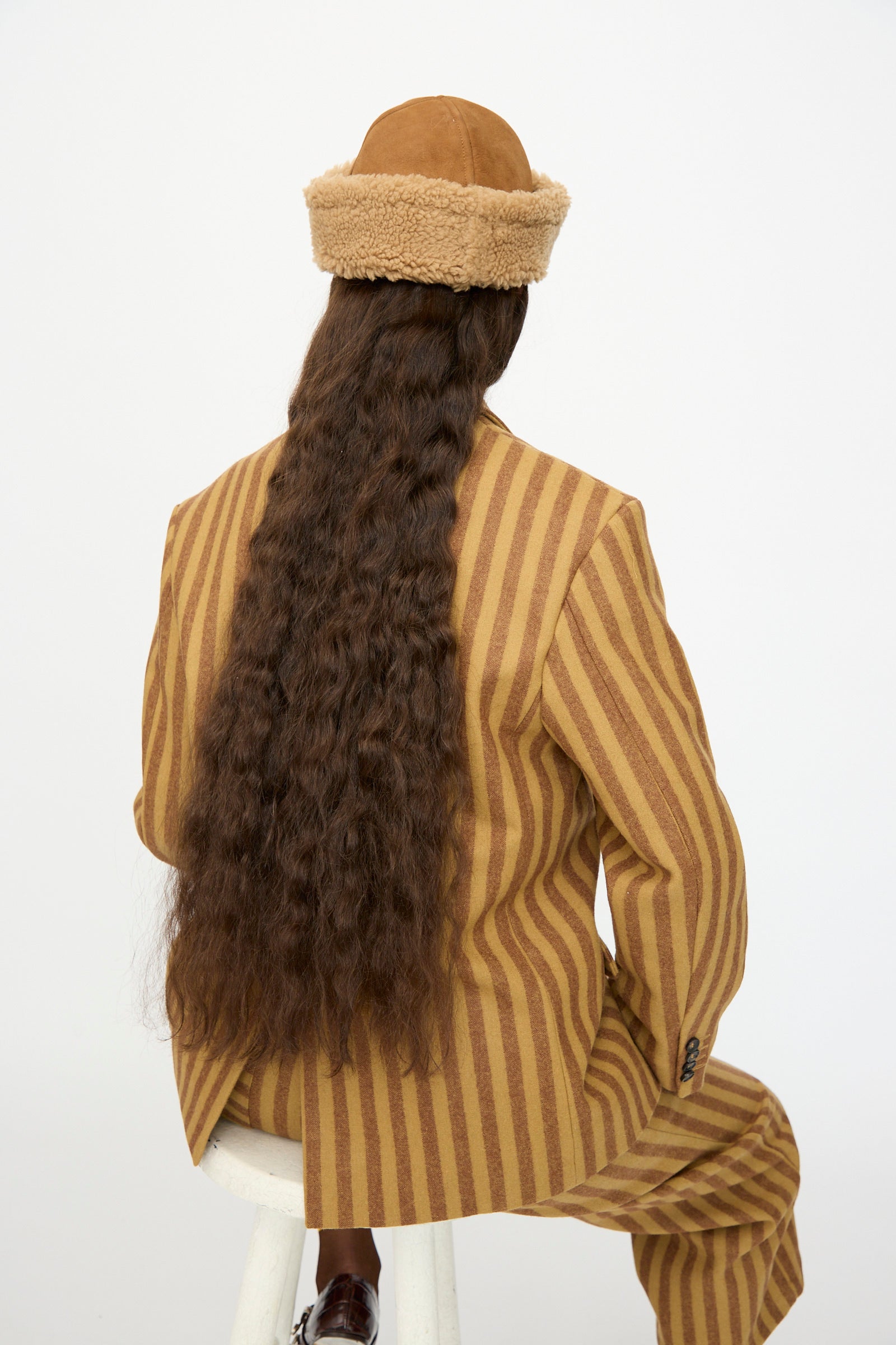 A person with long, wavy brown hair sits on a white stool, wearing a tan and brown striped suit and the Cawley Suede Back Curly Sheepskin Cap in Tan, facing away from the camera.