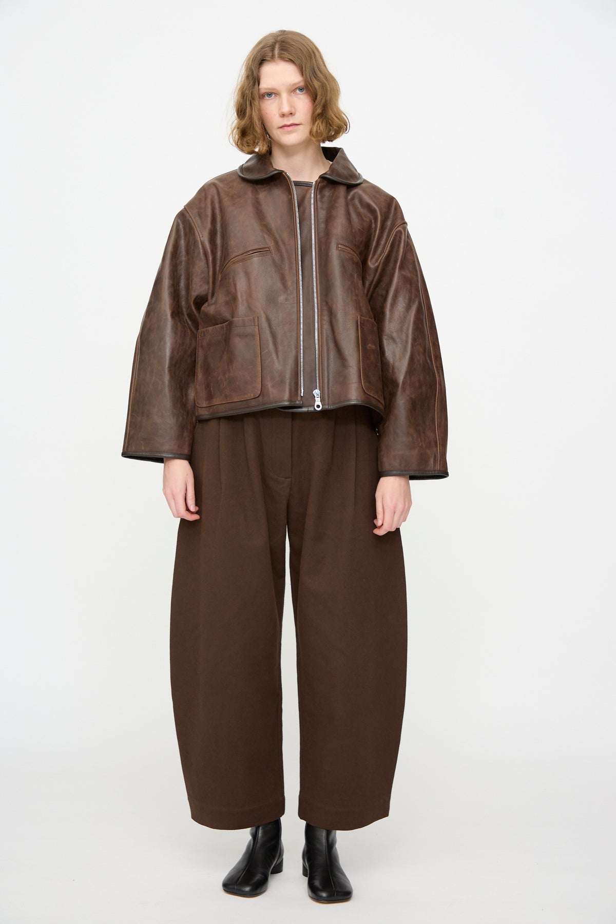 A person wears the Cawley Waxed Leather Flying Jacket in Walnut, paired with wide brown pants and black boots, standing against a plain white background.