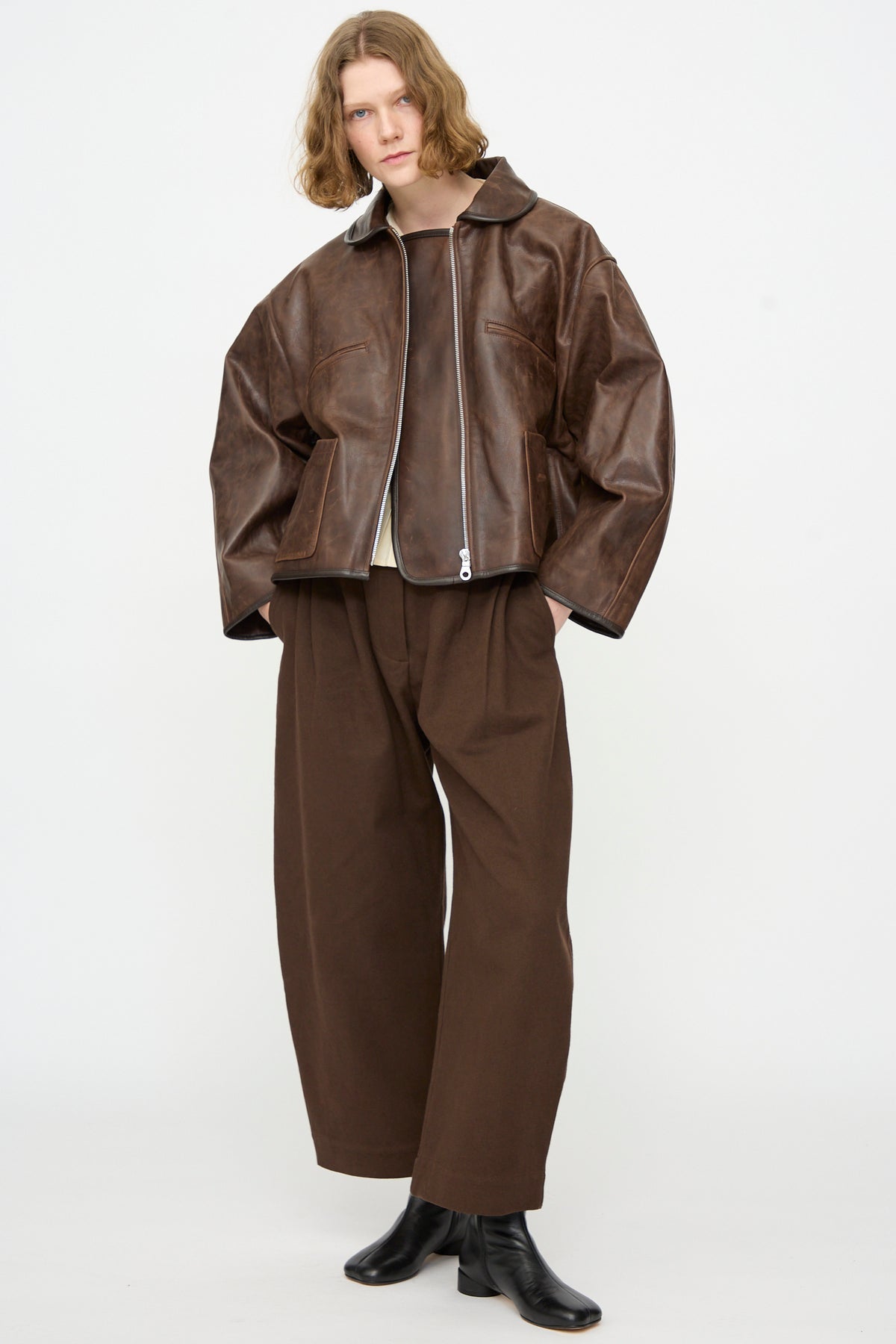 A person stands against a plain white background wearing the Cawley Waxed Leather Flying Jacket in Walnut, styled with wide brown pants, black boots, and a white shirt. The jacket’s oversized fit brings a modern edge to this classic look.