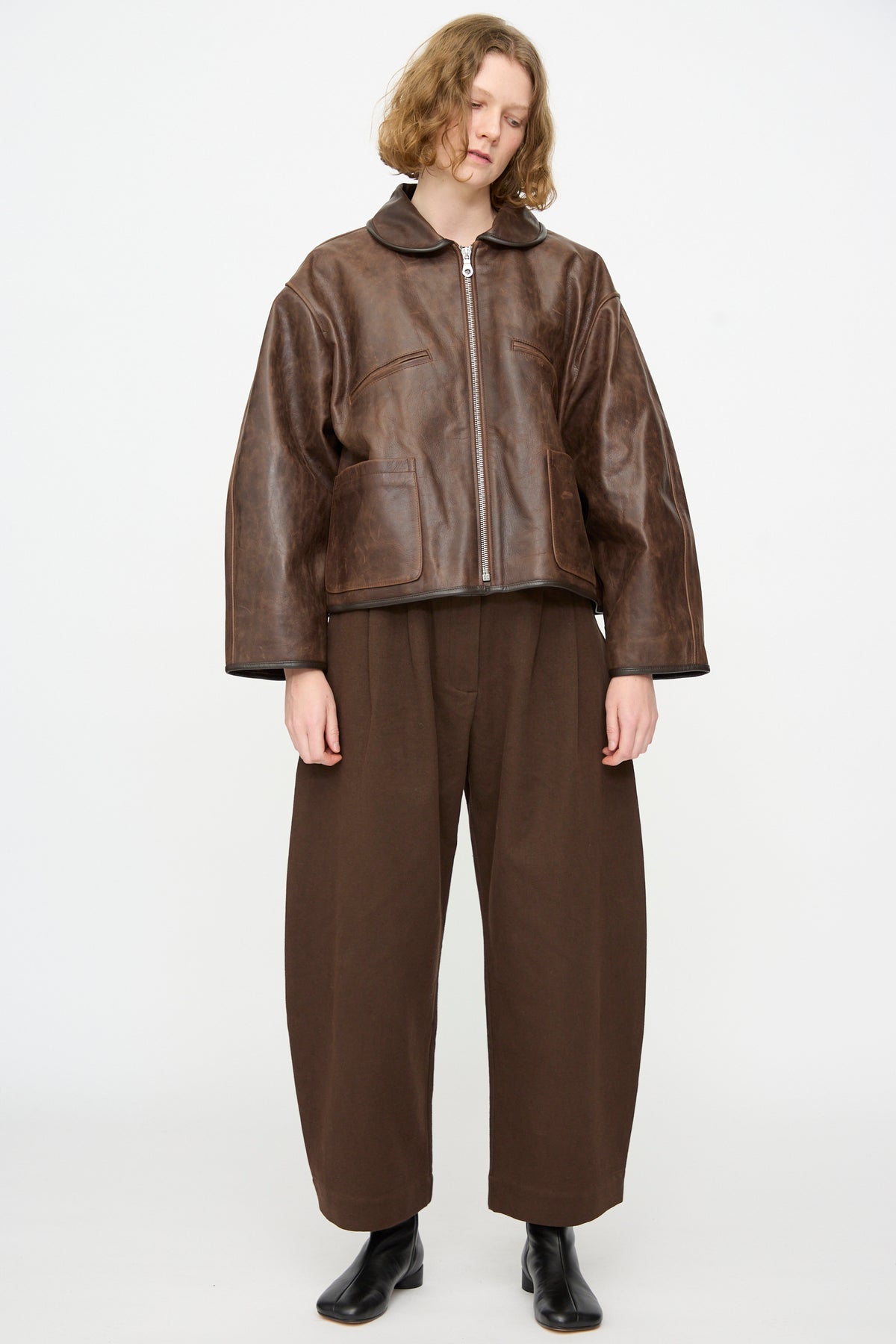 A person stands against a plain background wearing the Cawley Waxed Leather Flying Jacket in Walnut, styled with loose brown pants and black boots.