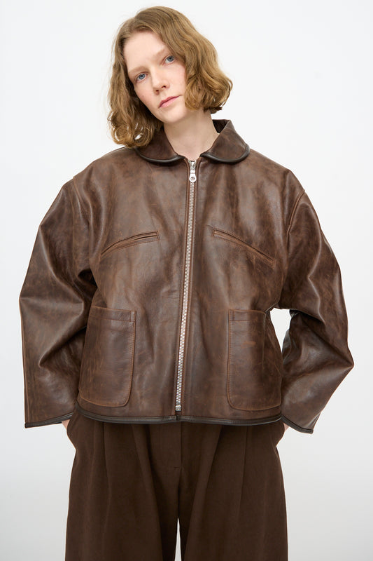 A person with shoulder-length light brown hair wears the Cawley Waxed Leather Flying Jacket in Walnut—an oversized fit aviator style featuring front pockets—paired with brown pants, standing against a plain white background.