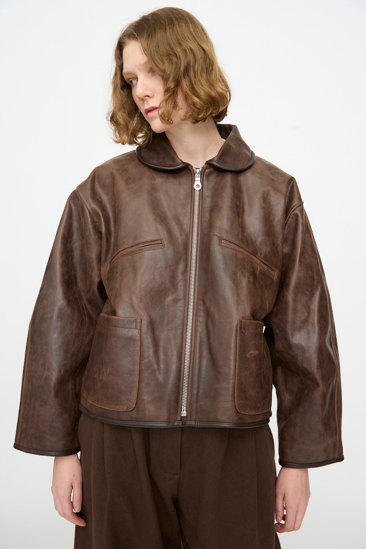 A person wearing the Cawley Waxed Leather Flying Jacket in Walnut, featuring a front zipper and large pockets, paired with brown pants and standing against a plain light background.
