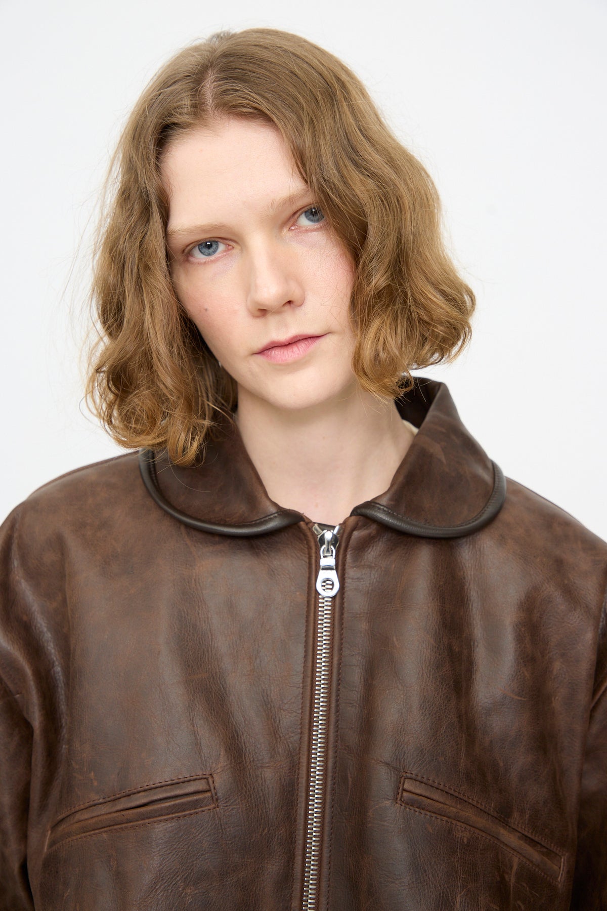A person with short, wavy light brown hair wears the Cawley Waxed Leather Flying Jacket in Walnut, featuring a zipper and rounded collar, standing against a plain white background.
