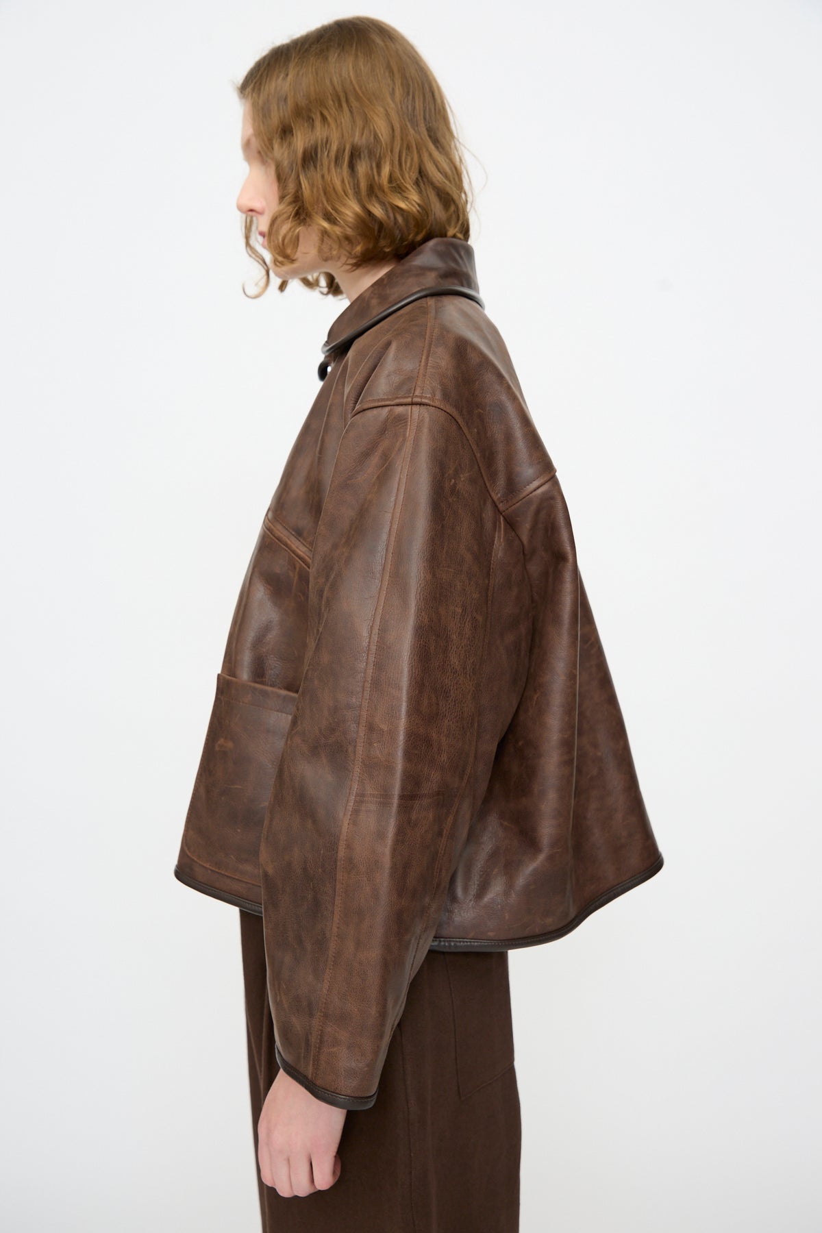 A person with medium-length wavy hair wearing the Cawley Waxed Leather Flying Jacket in Walnut stands in profile against a plain white background.