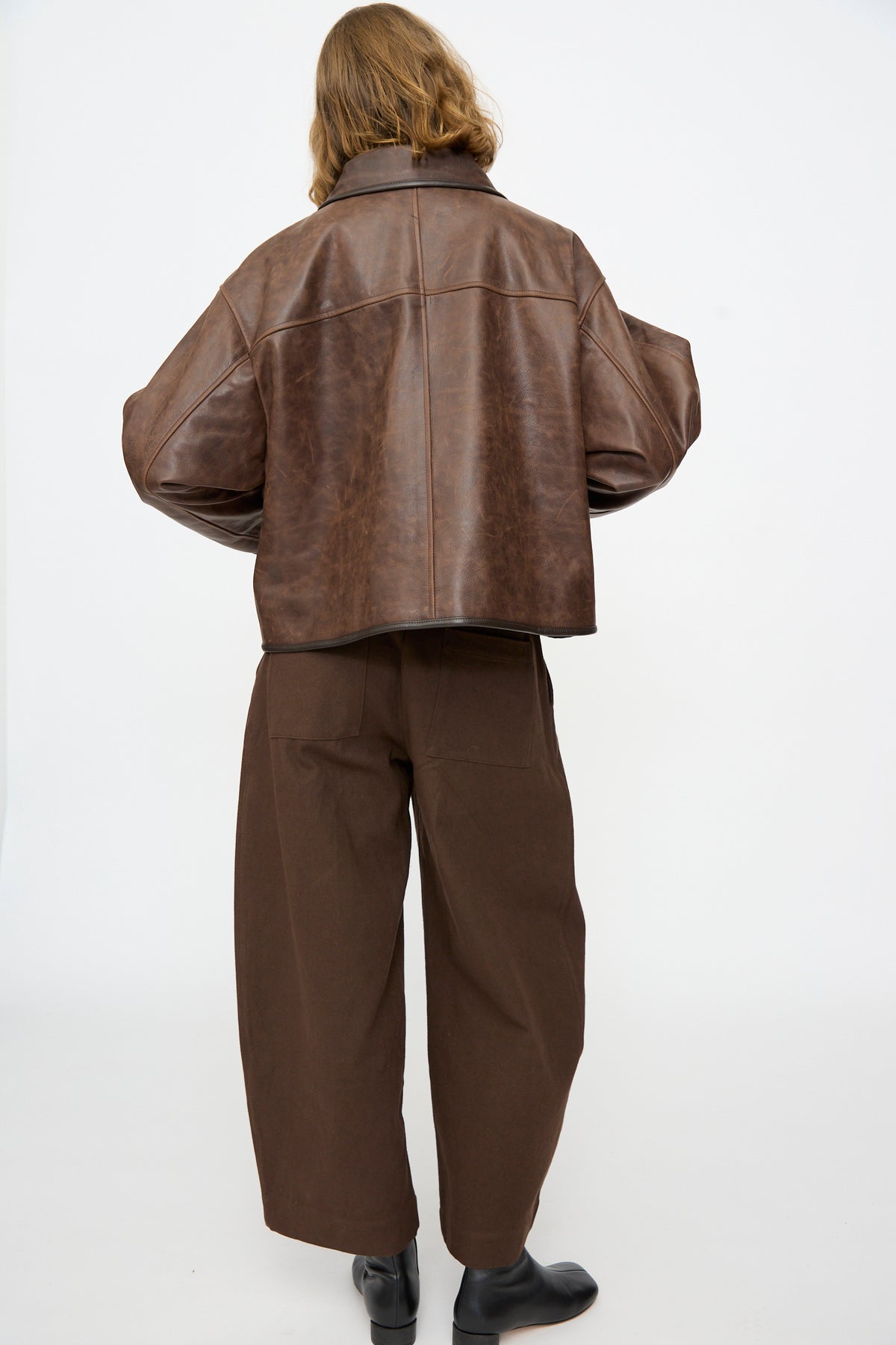 A person stands with their back to the camera, wearing the Cawley Waxed Leather Flying Jacket in Walnut—an oversized fit—paired with loose brown pants and black shoes, set against a plain white background.
