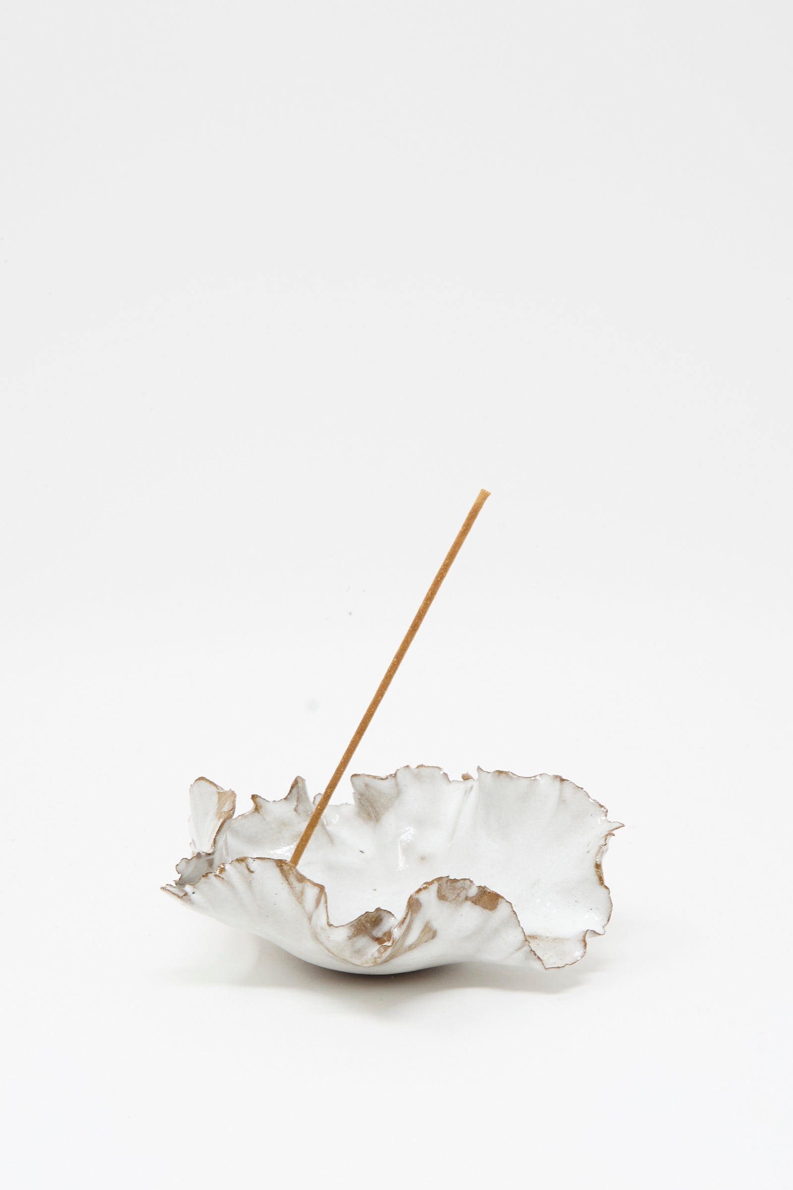 The Wild Petal Incense Holder by Chala Toprak is a white, ruffled stoneware piece featuring a single incense stick placed upright in its center, displayed against a plain white background.