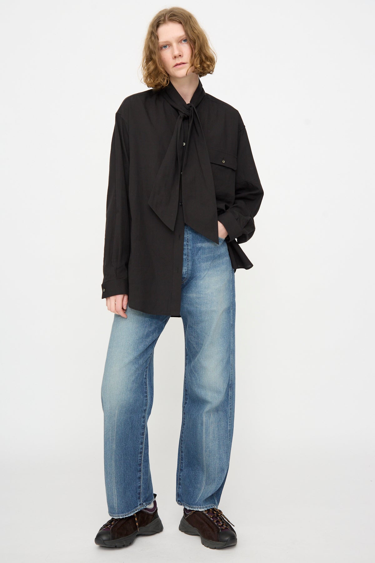 A person stands against a plain background wearing a loose black shirt with a large bow and the Chimala 12oz Denim Straight Cut in Work Light Blue, made in Japan.