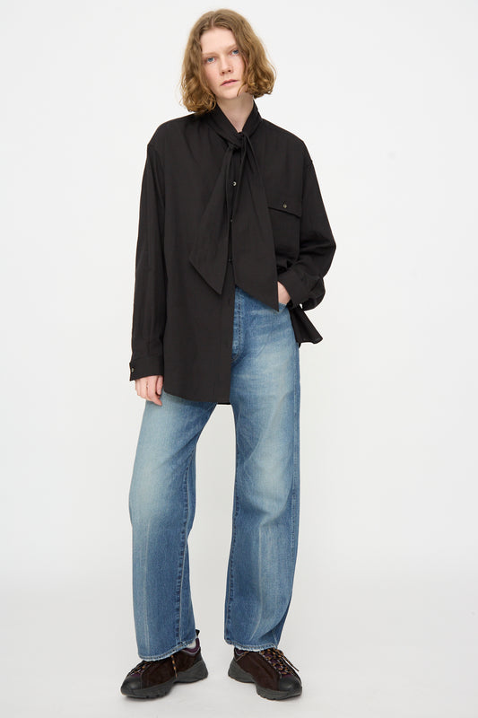 A person stands against a plain background wearing a loose black shirt with a large bow and the Chimala 12oz Denim Straight Cut in Work Light Blue, made in Japan.
