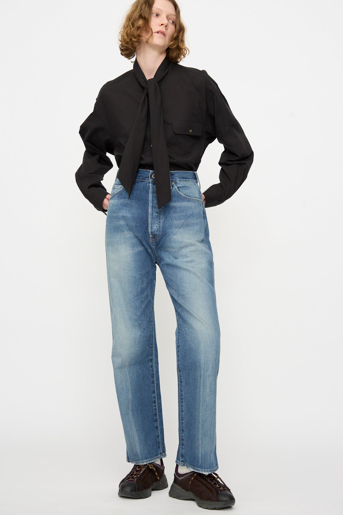 Person standing against a white background, wearing a black long-sleeve shirt with a tie, Chimala 12oz Denim Straight Cut in Work Light Blue high-waisted selvedge jeans made in Japan, and dark shoes.