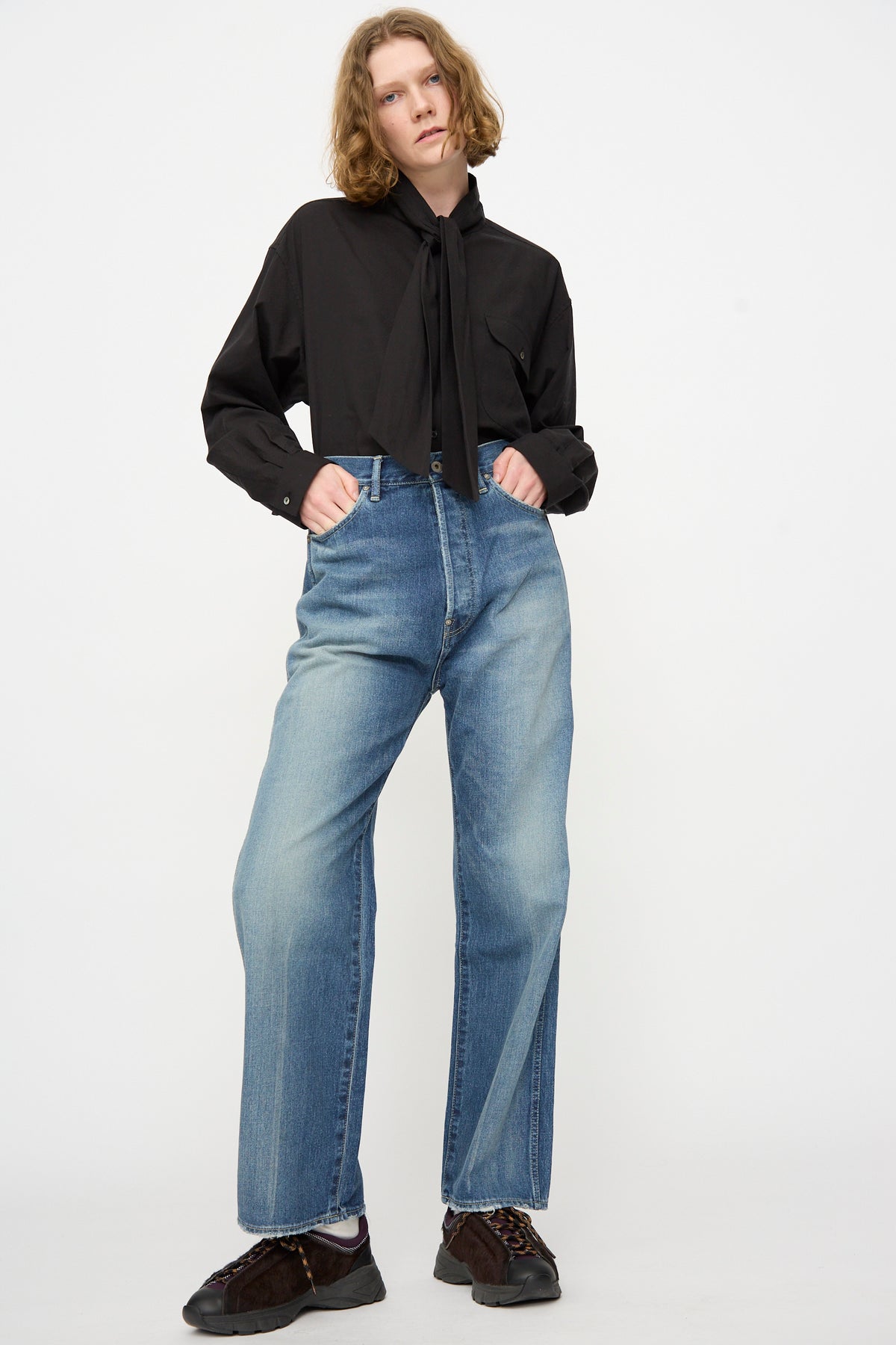 A person stands against a white background, wearing a black long-sleeve shirt with a tie detail and Chimala 12oz Denim Straight Cut jeans in Work Light Blue, hands in pockets, paired with brown shoes.