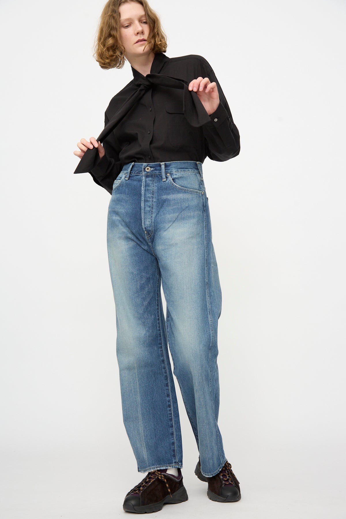 A person stands against a plain background, wearing a black button-up shirt, Chimala’s 12oz Denim Straight Cut in Work Light Blue jeans with a high waist and straight pant style, along with dark shoes. They hold one side of their shirt collar and gaze slightly away.