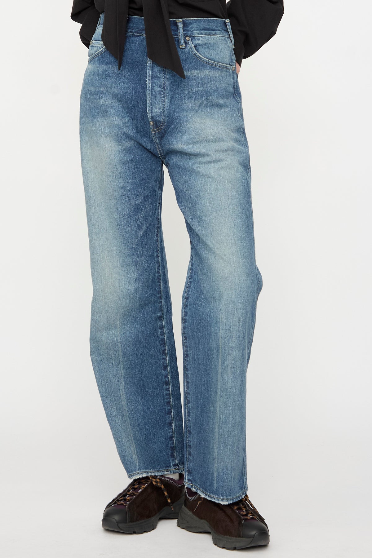 A person is wearing Chimala’s 12oz Denim Straight Cut in Work Light Blue jeans, paired with a black top and black shoes with brown detail, standing against a plain background.