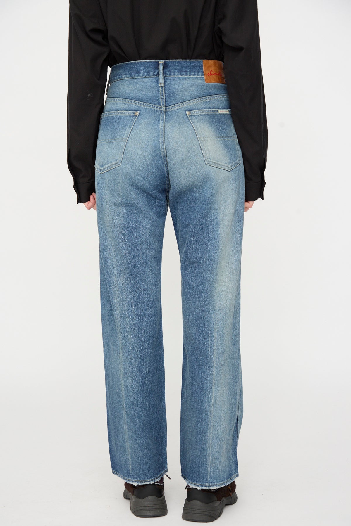 A person wearing Chimala’s 12oz Denim Straight Cut in Work Light Blue, paired with a black long-sleeve shirt and black shoes, seen from the back against a plain white background.
