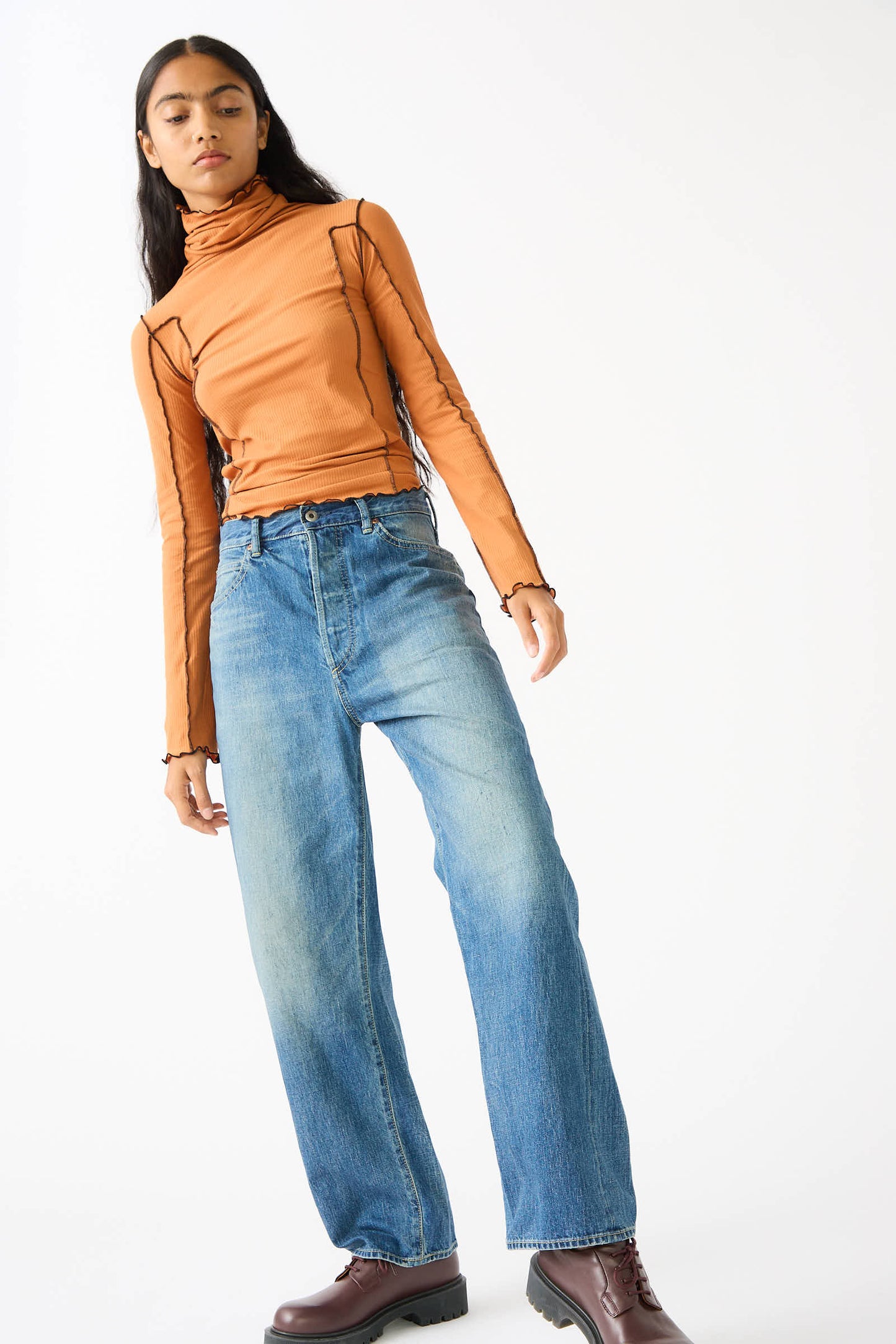 A person stands wearing a brown turtleneck, Chimala's 12 oz. Selvedge Denim Monroe Cut in Medium Wash jeans with a relaxed fit, and brown boots, against a plain white background.