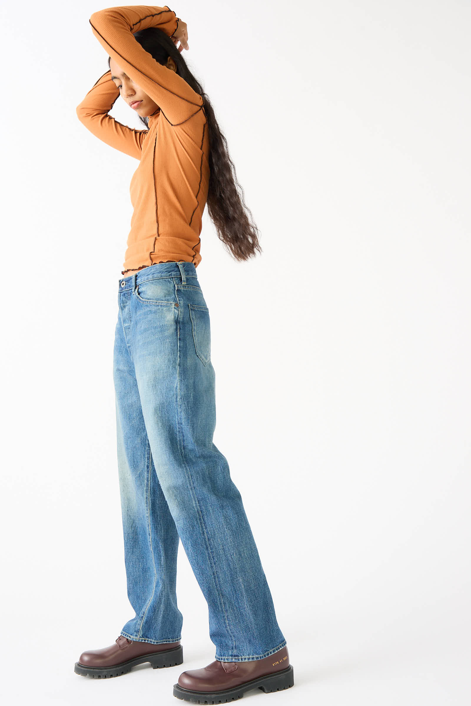 A person stands in a relaxed pose, wearing a long-sleeved orange top, Chimala's 12 oz. Selvedge Denim Monroe Cut in Medium Wash blue jeans, and brown shoes, with long hair flowing down their back.