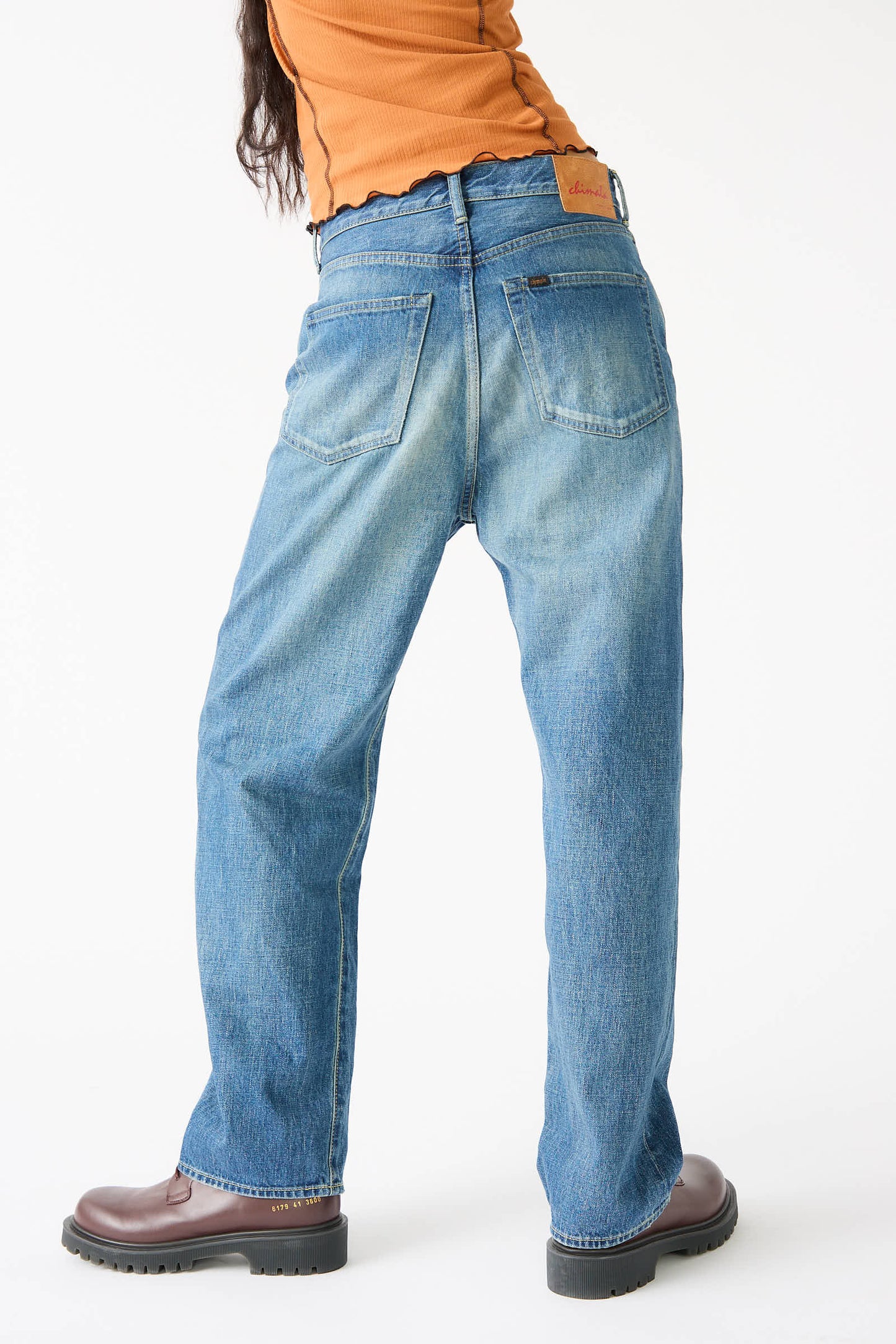 A person wearing Chimala's 12 oz. Selvedge Denim Monroe Cut in Medium Wash, a rust-colored top, and brown boots stands with their back facing the camera against a plain white background.