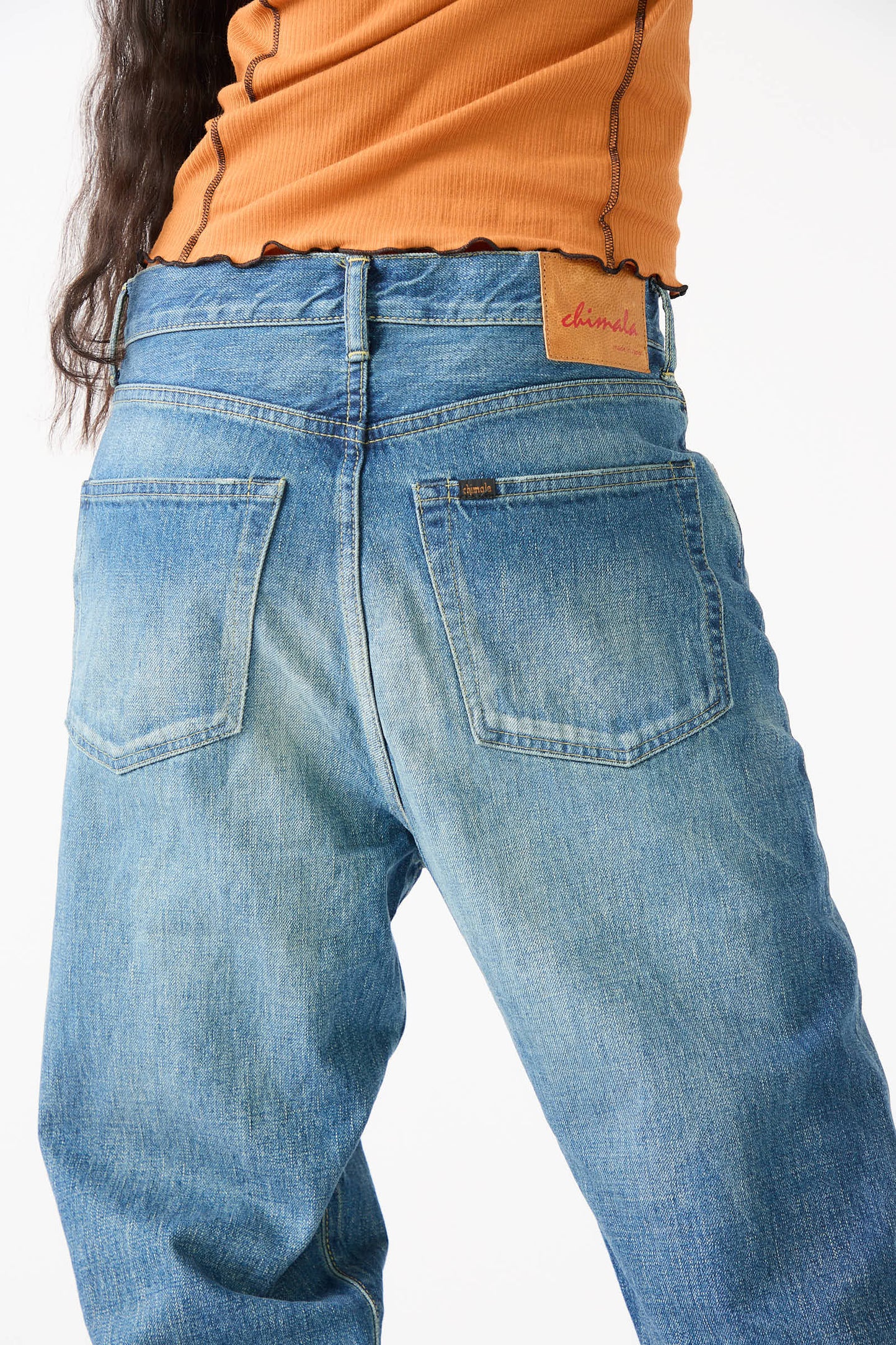 Person wearing 12 oz. Selvedge Denim Monroe Cut in Medium Wash by Chimala and an orange top, showing the back view with focus on the high-waisted pant pockets and waistband.