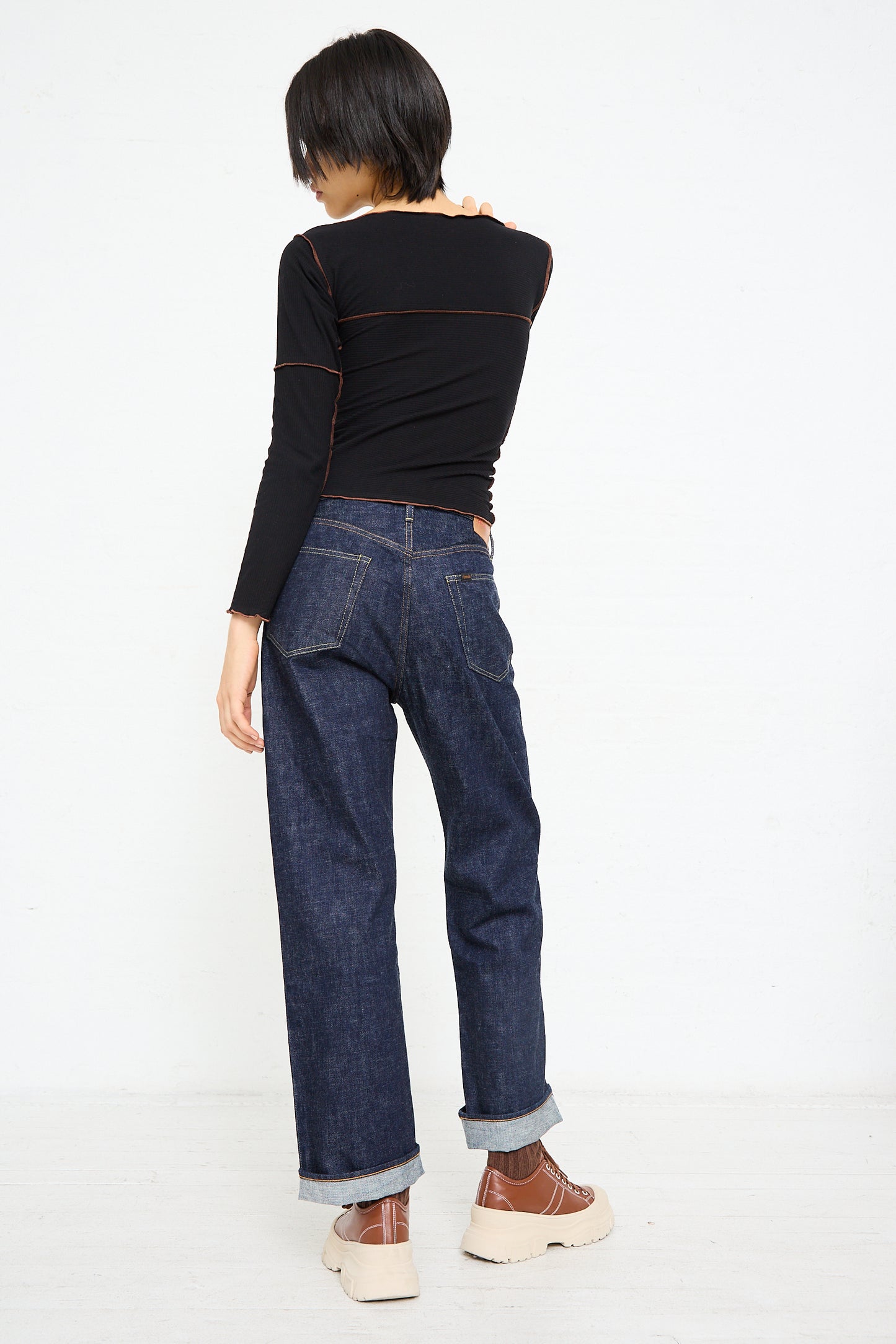 A person standing with their back to the camera, wearing a black top and blue denim Chimala 13.5 oz. Selvedge Denim Straight Cut in Rinse jeans, with one hand reaching across to touch the opposite.