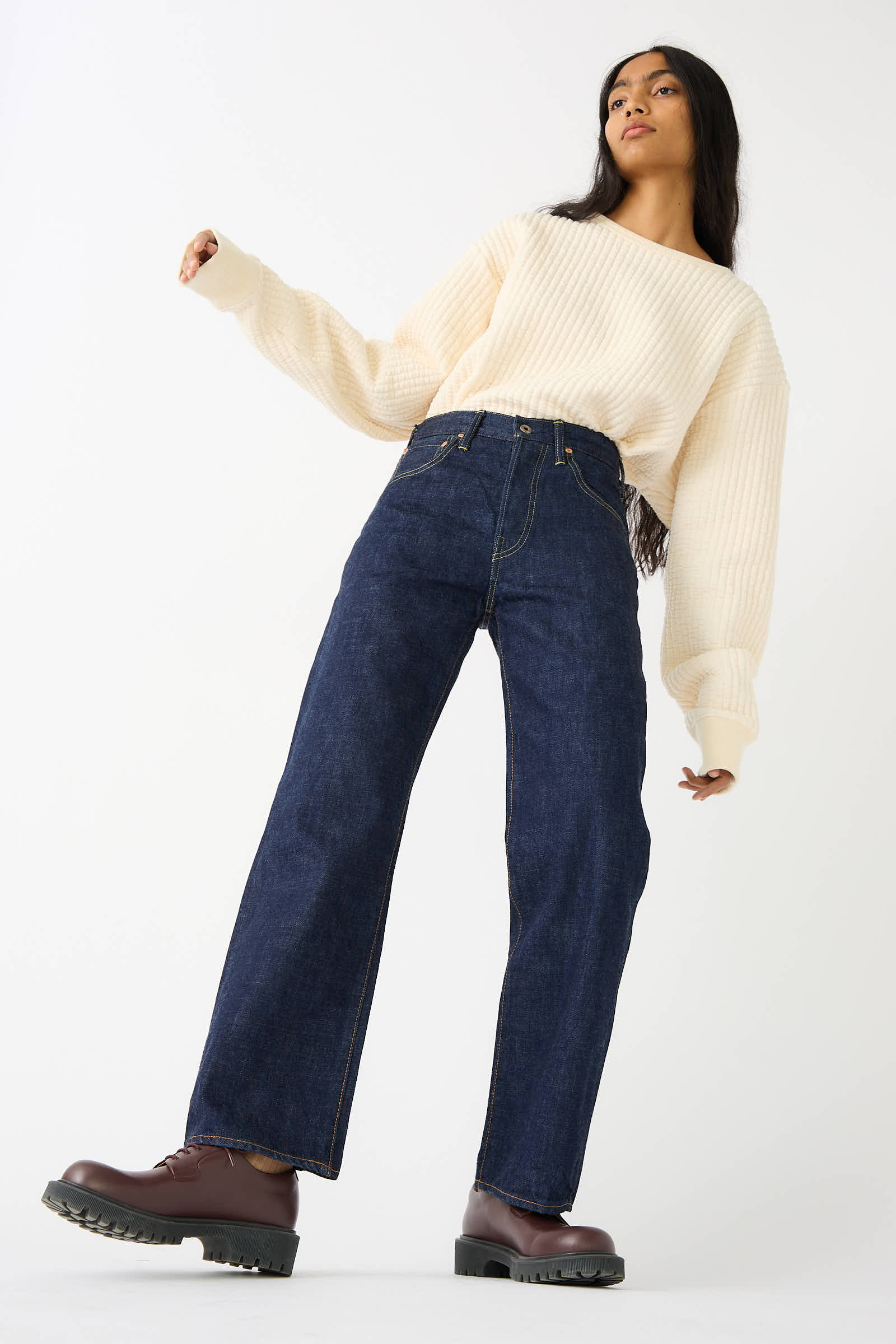 A person with long hair wears a cream-colored sweater, Chimala's 13.5 oz Selvedge Denim Straight Cut in Rinse, and brown boots, standing with one arm bent and the other relaxed against a plain light background.