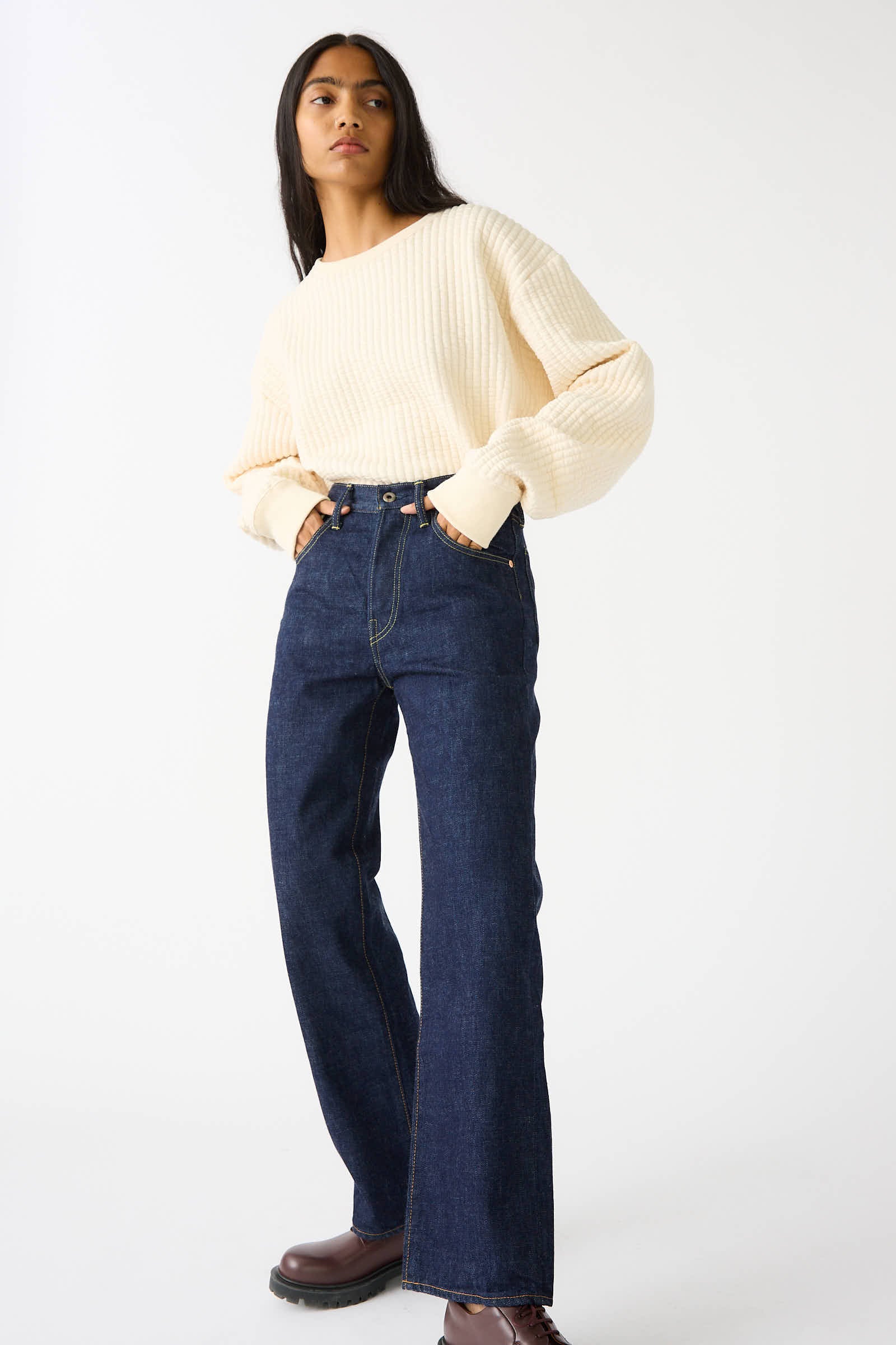 A person with long dark hair wears a cream sweater, Chimala's 13.5 oz Selvedge Denim Straight Cut in Rinse, and brown shoes. The relaxed fit ensemble exudes effortless style as they stand with hands in pockets against a plain white background.