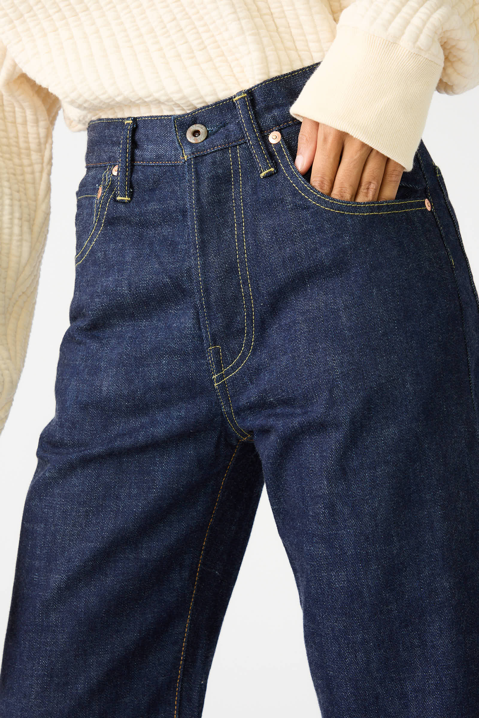 A person wearing a cream-colored ribbed sweatshirt made from 100% cotton, with the sleeve partially covering their hand, which is placed in the front pocket of relaxed fit dark blue jeans from Chimala's 13.5 oz Selvedge Denim Straight Cut in Rinse collection.