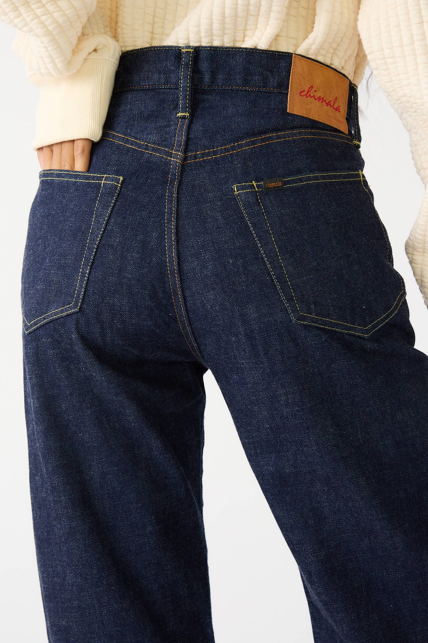 A person wearing Chimala's 13.5 oz Selvedge Denim Straight Cut in Rinse, high-waisted dark blue jeans made from 100% cotton, featuring a relaxed fit with a brown leather patch on the waistband.