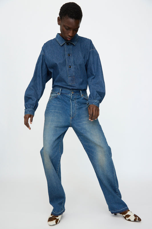 Someone wearing a blue denim button-up shirt, Chimala 13oz. Selvedge Denim Monroe Cut in Dark Distress jeans, and cow-print shoes stands against a plain white background.