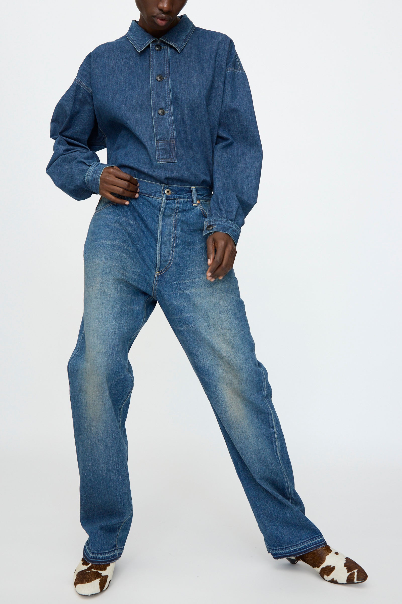 A person wears Chimala’s 13oz. Selvedge Denim Monroe Cut in Dark Distress jeans with a blue denim button-up and brown-and-white cow-print shoes, standing against a plain background.