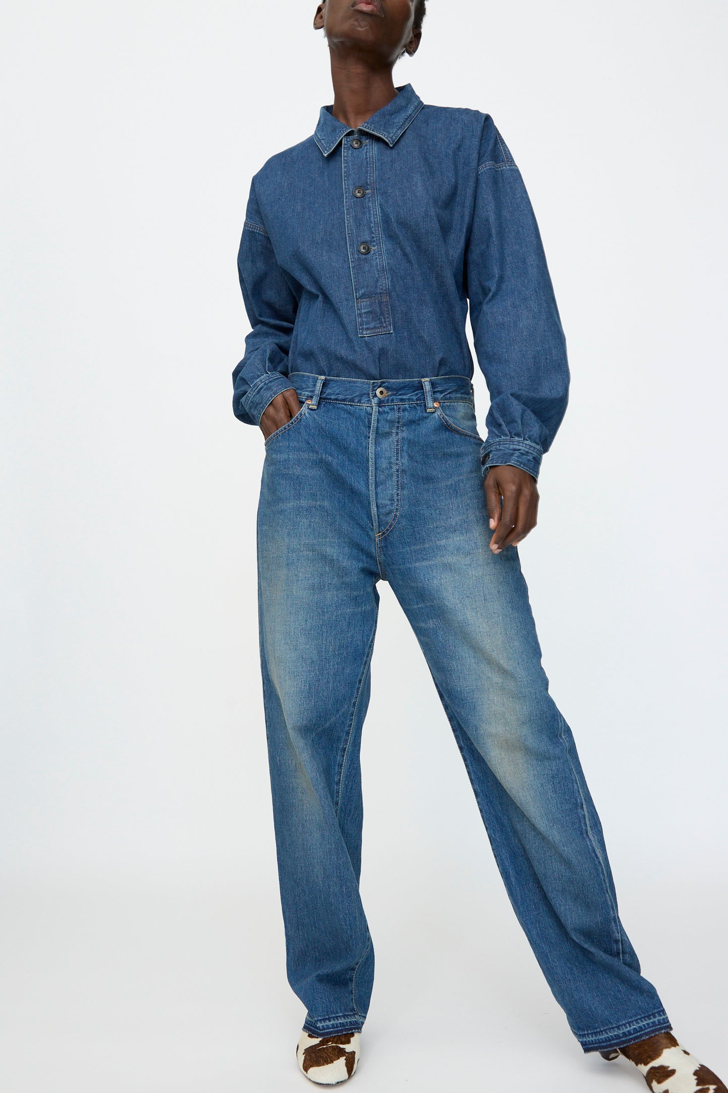 A person wears Chimala's 13oz. Selvedge Denim Monroe Cut in Dark Distress jeans, paired with a long-sleeve denim shirt, standing with one hand in a pocket against a plain light background.