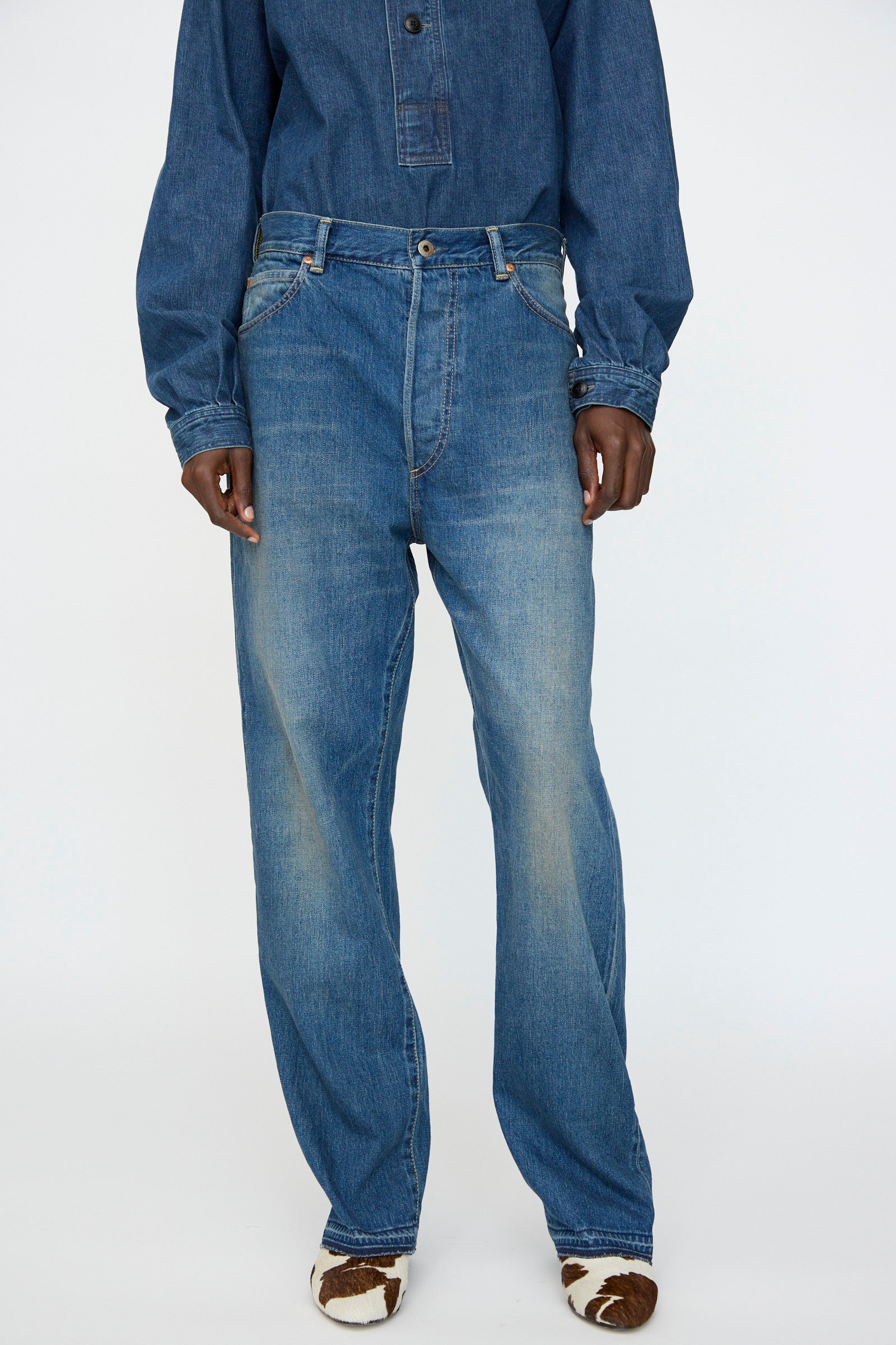 A person wearing Chimala’s 13oz. Selvedge Denim Monroe Cut in Dark Distress, paired with a blue denim top and brown-and-white cow-print shoes, stands against a plain light background.